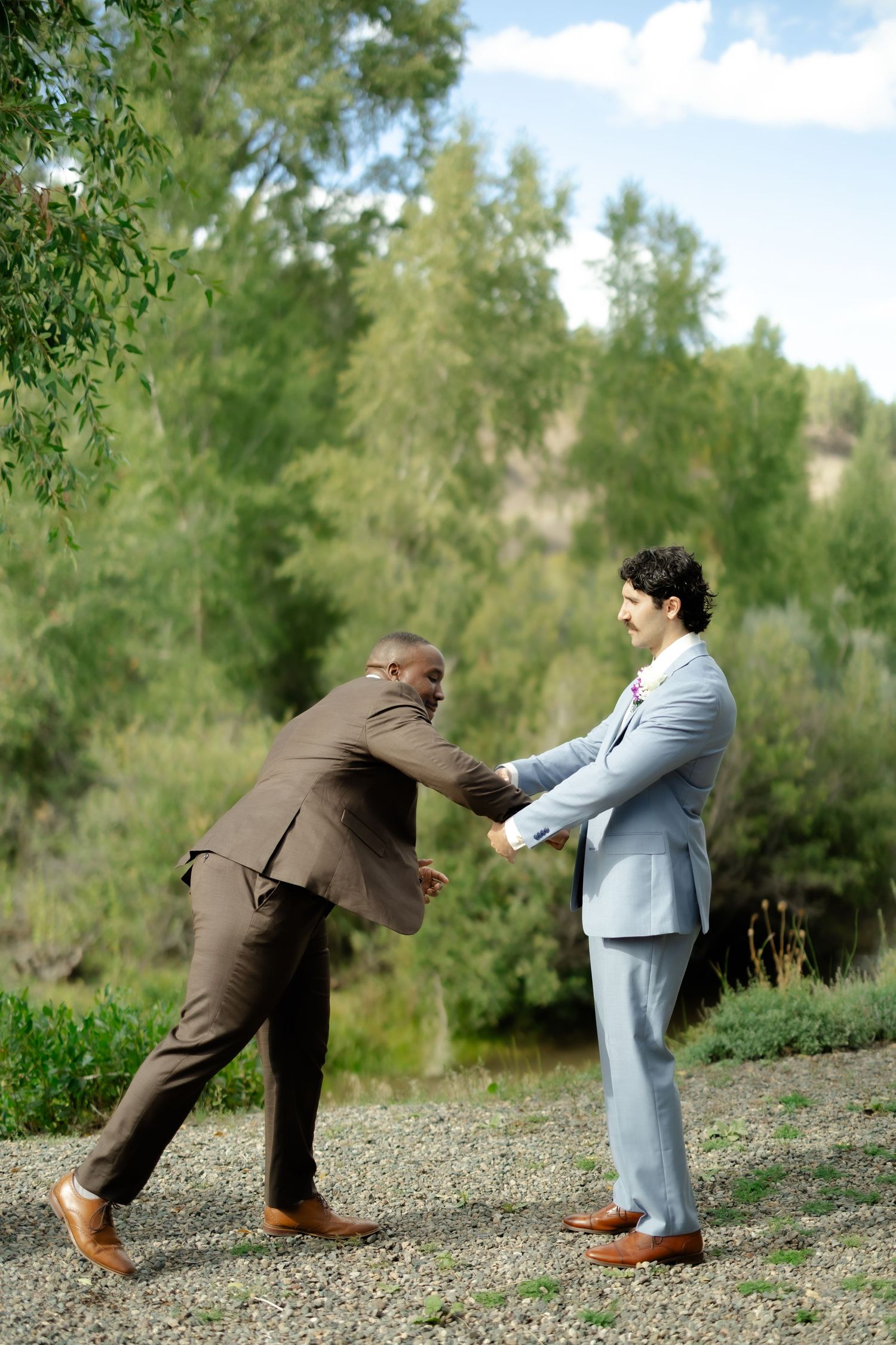 Two grooms in suits laughing and holding hands outdoors at a wedding ceremony with lush green trees behind them.