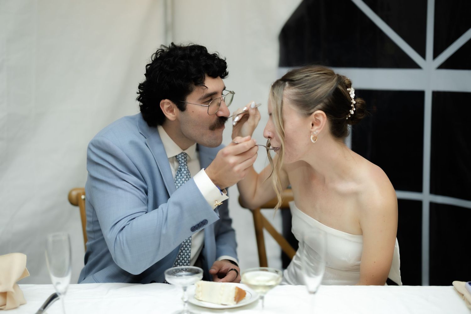 Bride and groom feeding each other wedding cake at reception table, wearing white dress and blue suit.