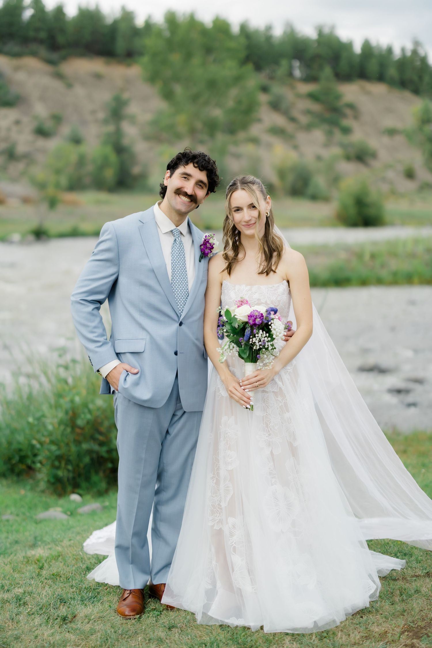 Bride and groom posing outdoors near a river, groom in light blue suit, bride in floral wedding gown holding purple bouquet.