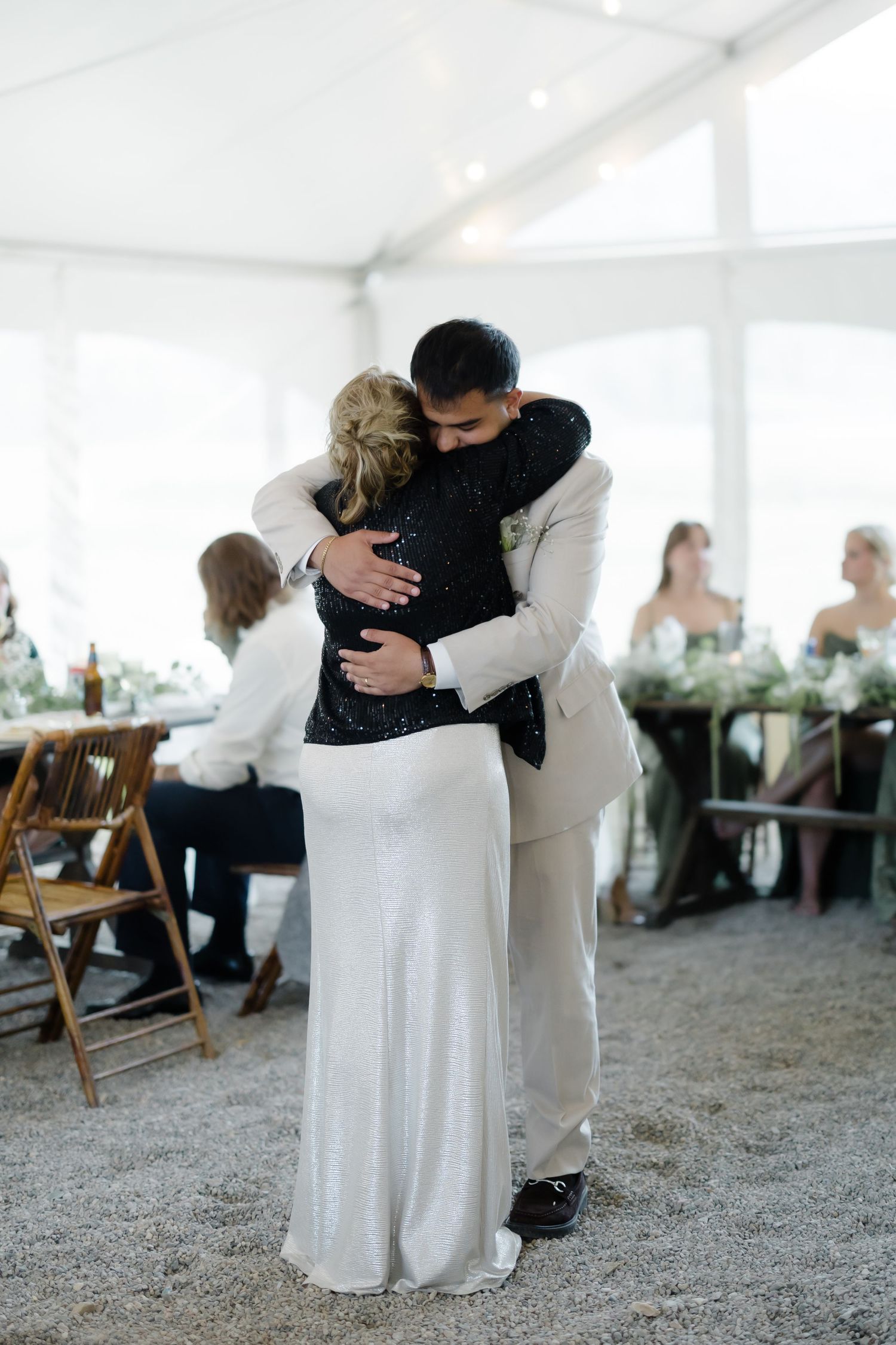 Groom in white suit embracing mother in black sequin dress during wedding reception dance.
