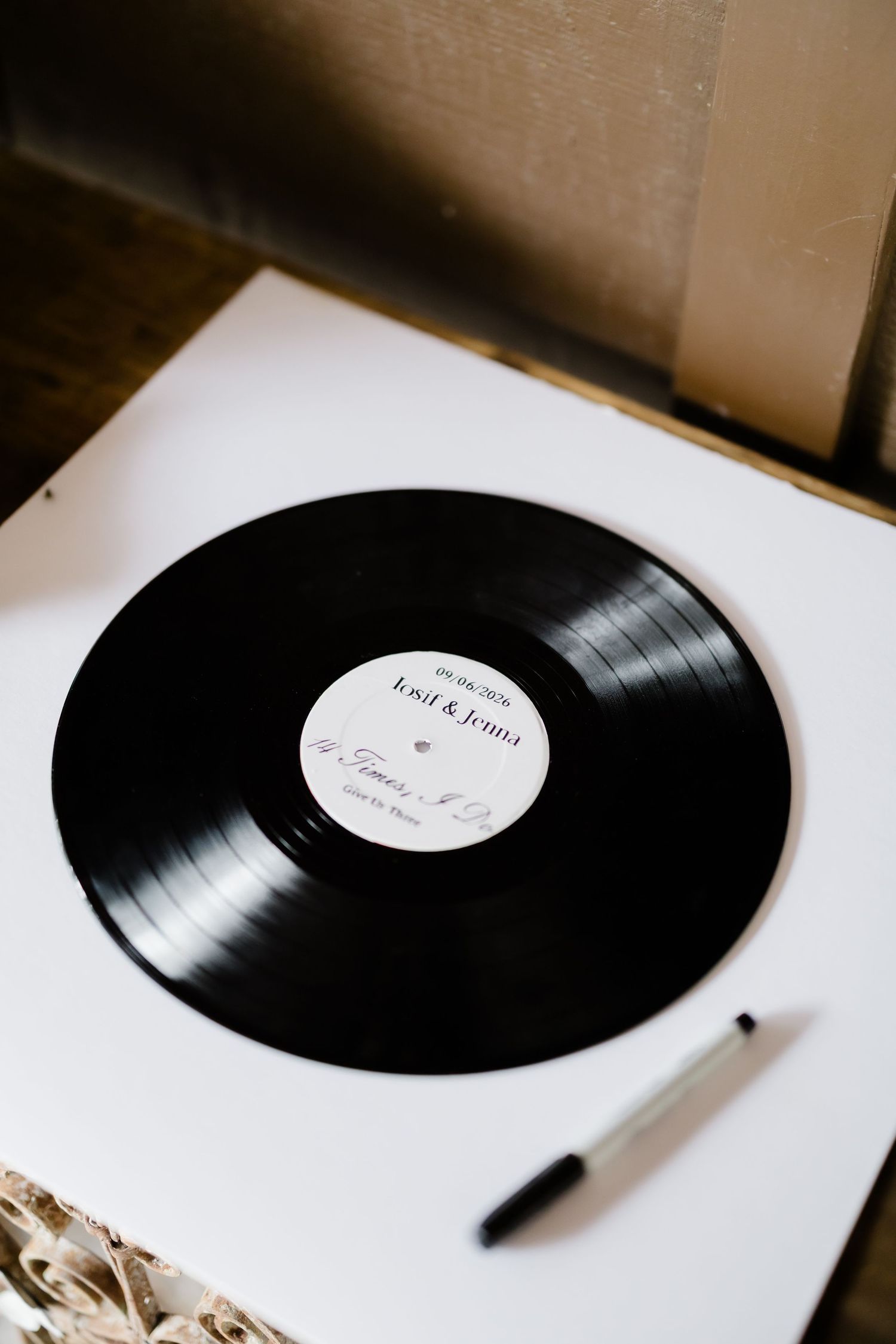 Black vinyl record wedding guest book with couple's names on label, pen beside it on white surface.