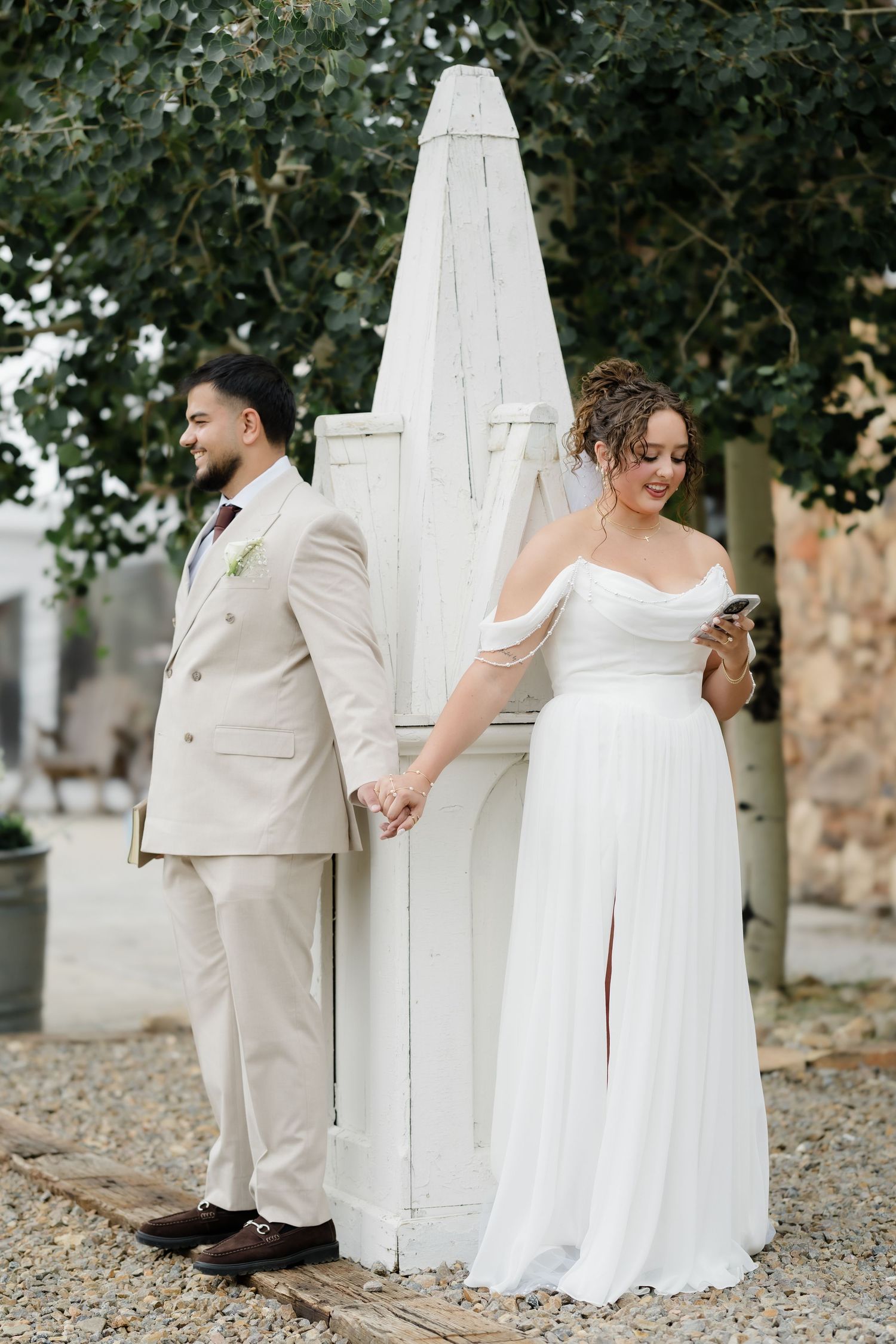 A bride in a white off-shoulder gown holds hands with a groom in a beige suit near a white wooden structure outdoors.