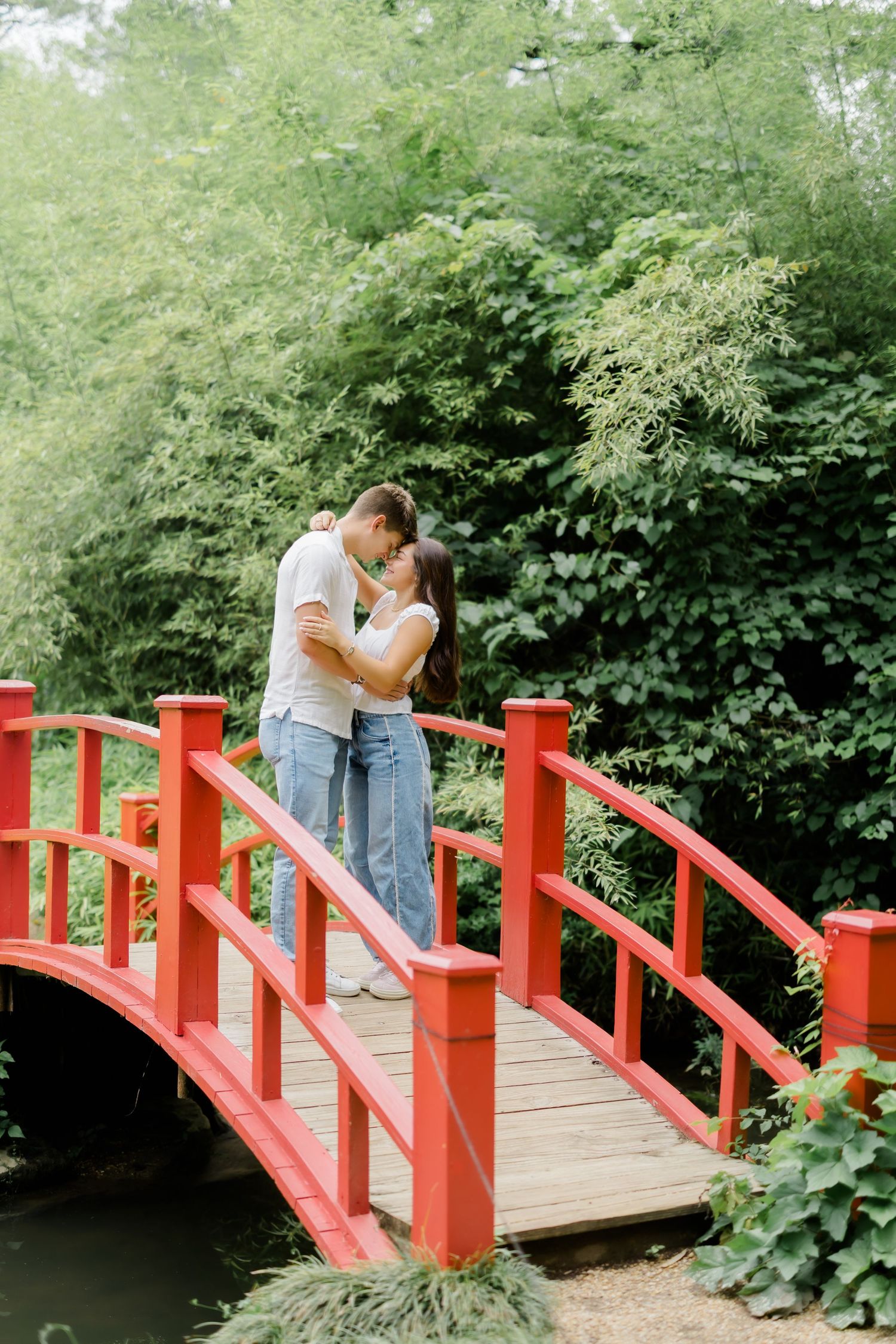Couple sharing a romantic kiss on a red arched bridge surrounded by lush green foliage in a garden.