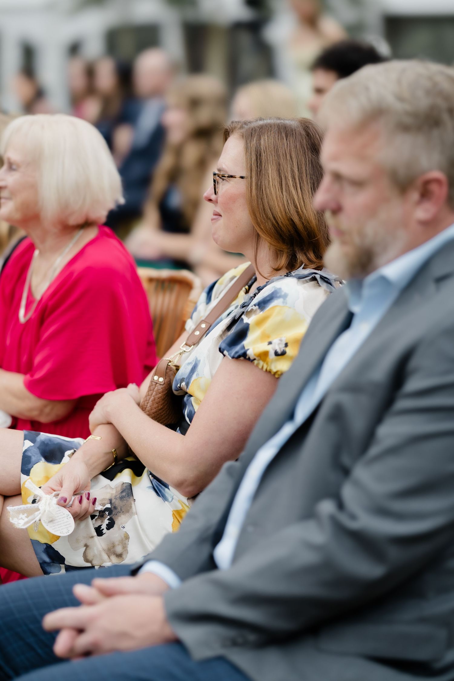 Wedding guests seated outdoors smiling and enjoying the ceremony, woman in floral dress holding a cup.