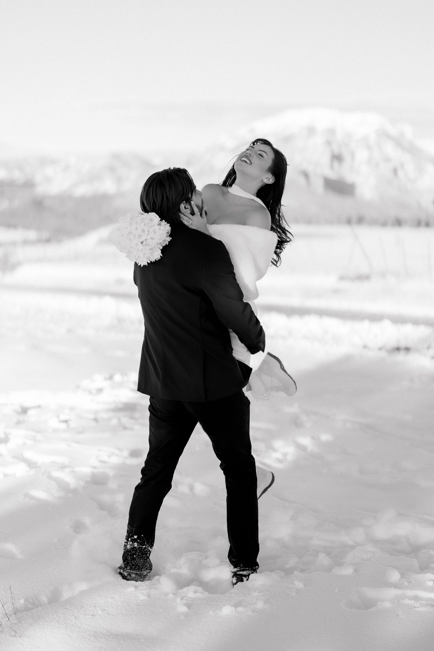Groom lifts bride in white dress as she laughs joyfully against snowy mountain landscape backdrop.