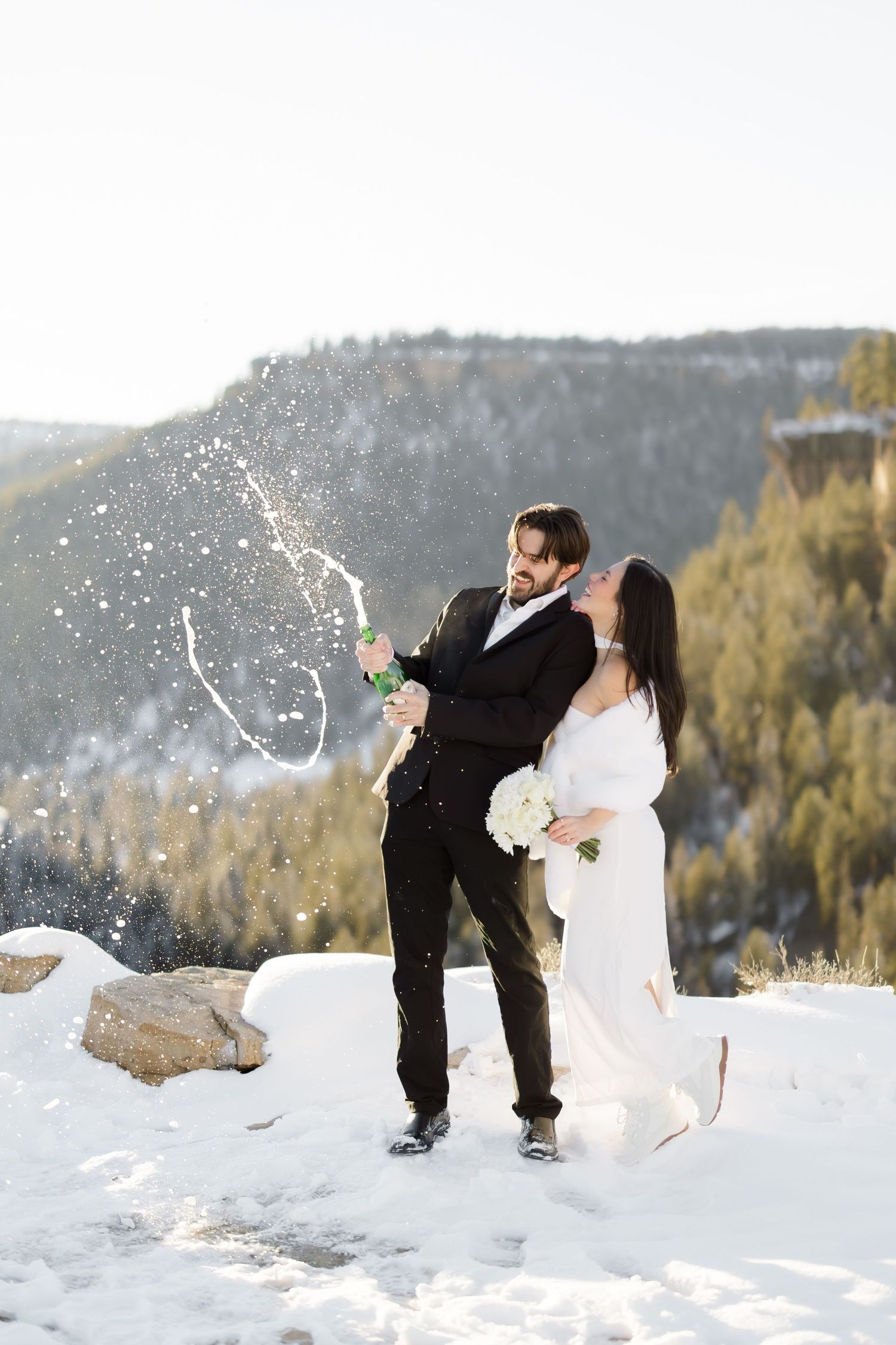 Wedding couple pops champagne bottle in snowy mountain landscape, bride holds white bouquet.