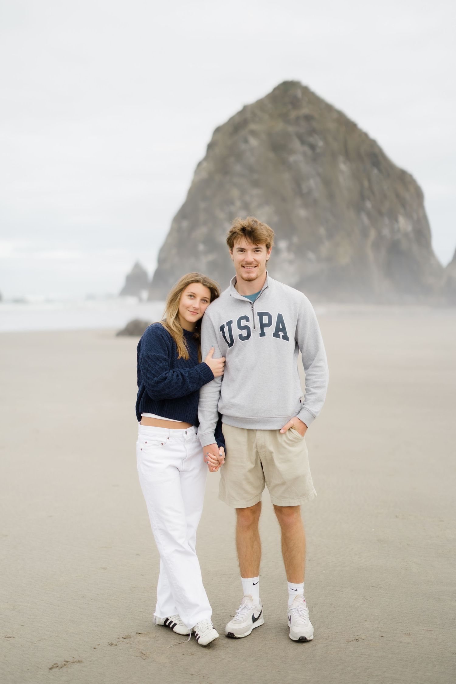 A couple smiles on a sandy beach near Haystack Rock at Cannon Beach, Oregon, wearing casual coastal attire.