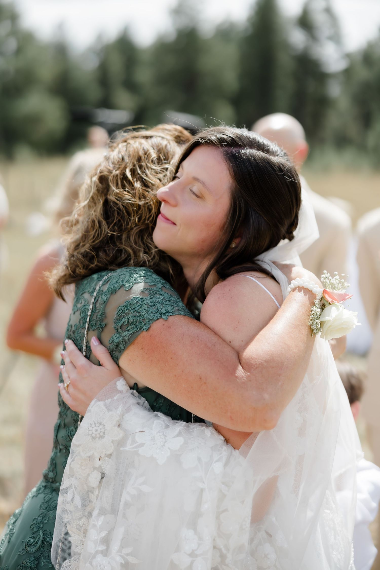 Bride in white lace gown shares emotional hug with guest in green lace dress at outdoor wedding ceremony.