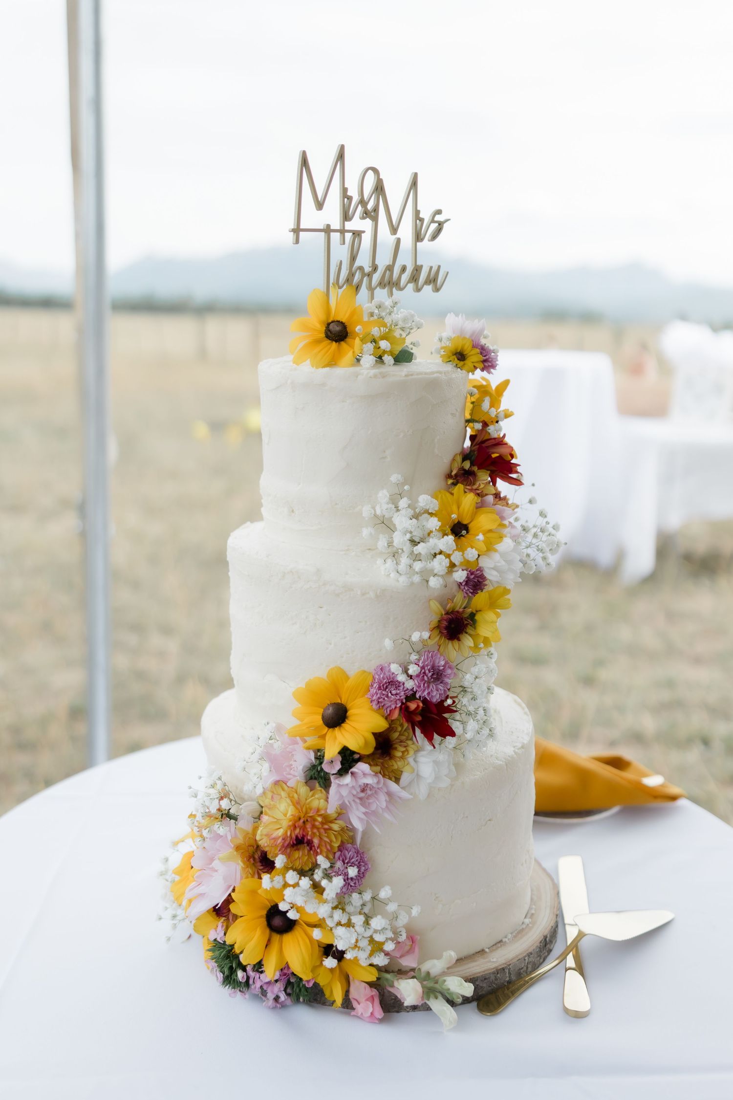 Three-tier white wedding cake with sunflowers and wildflowers, Mr & Mrs Thibdeau topper, displayed outdoors at rustic wedding.