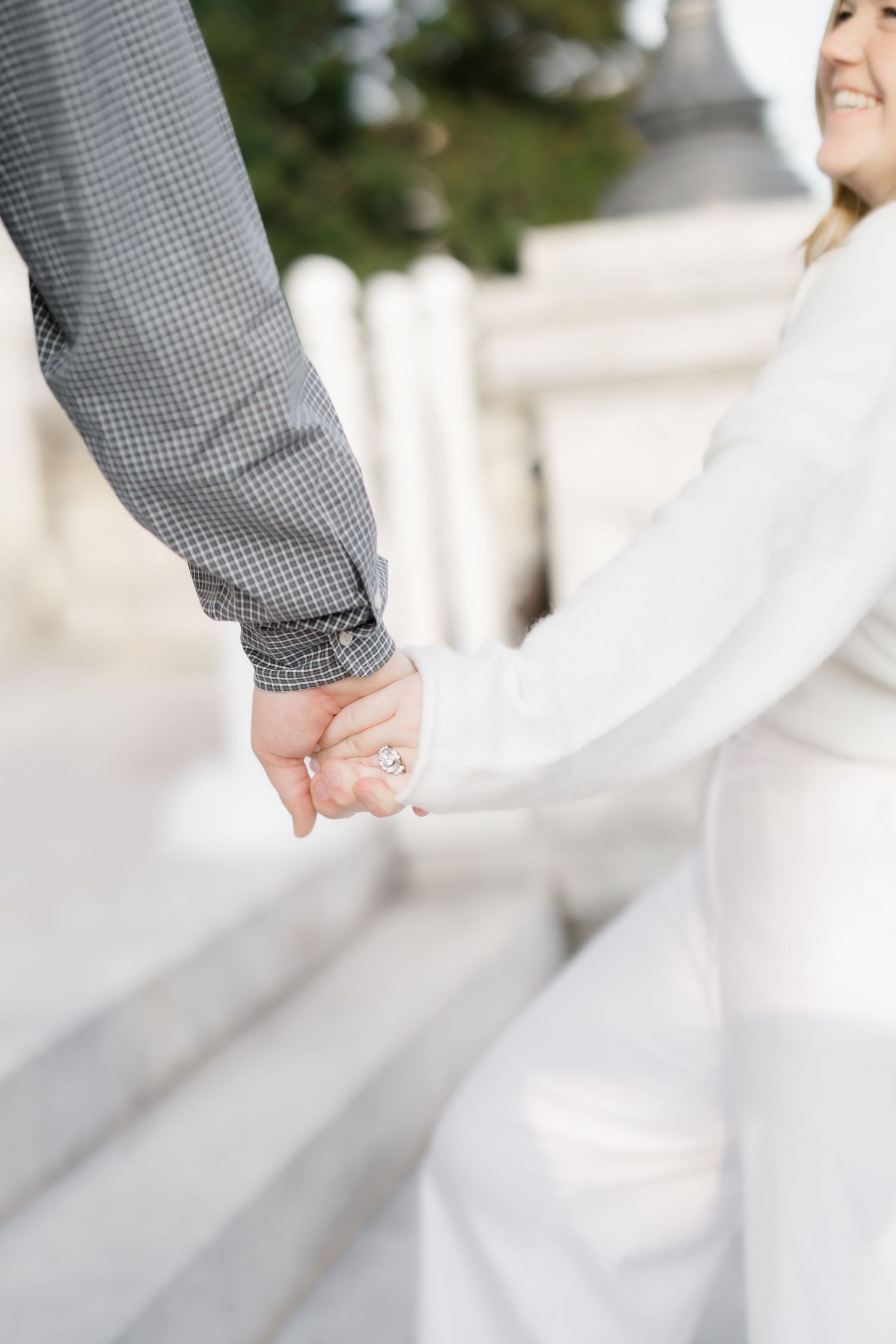 Couple holding hands on white marble steps, bride wearing engagement ring in white dress, groom in gray checkered suit.