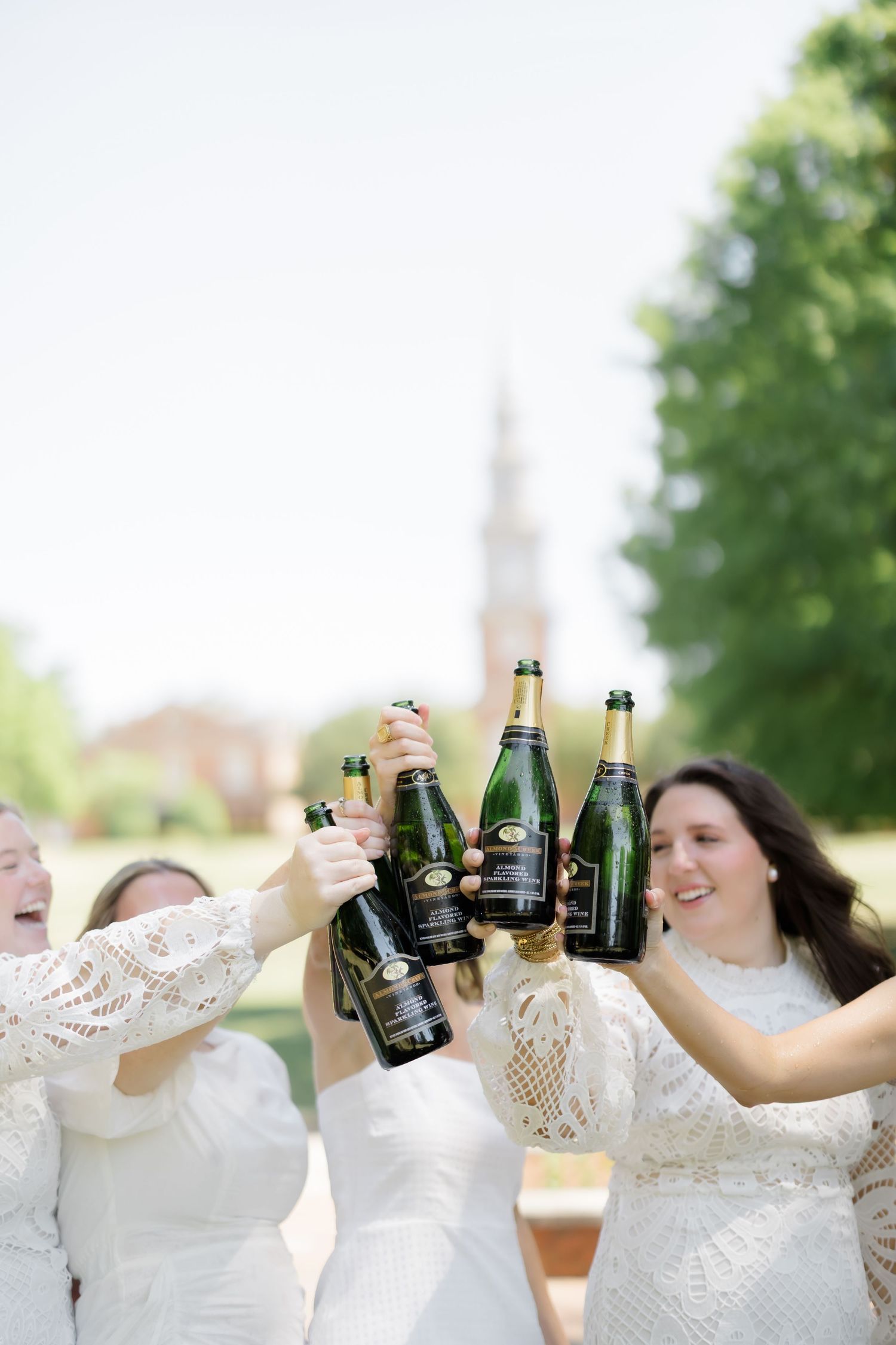 Bridesmaids in white dresses toasting with champagne bottles outdoors near a brick church on a sunny day.
