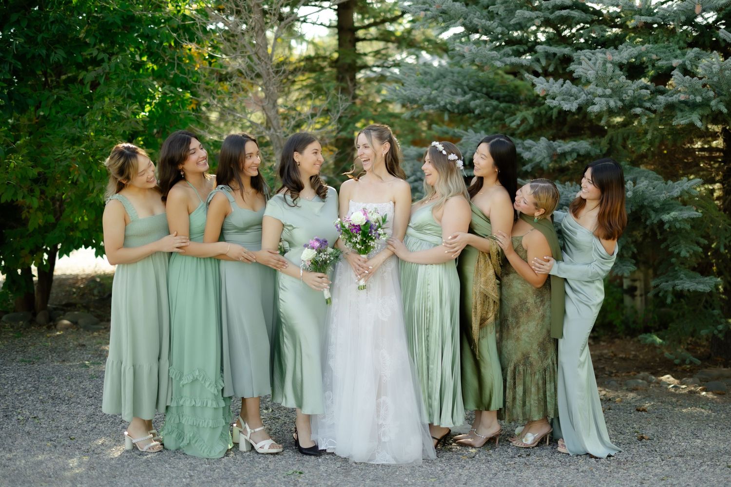 Bride in white gown with bridesmaids in sage green dresses laughing outdoors among pine trees.