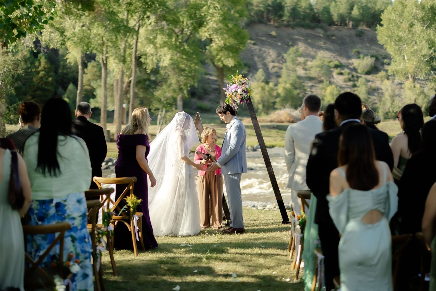 Outdoor wedding ceremony on green lawn with bride and groom at floral arch, guests seated among trees.