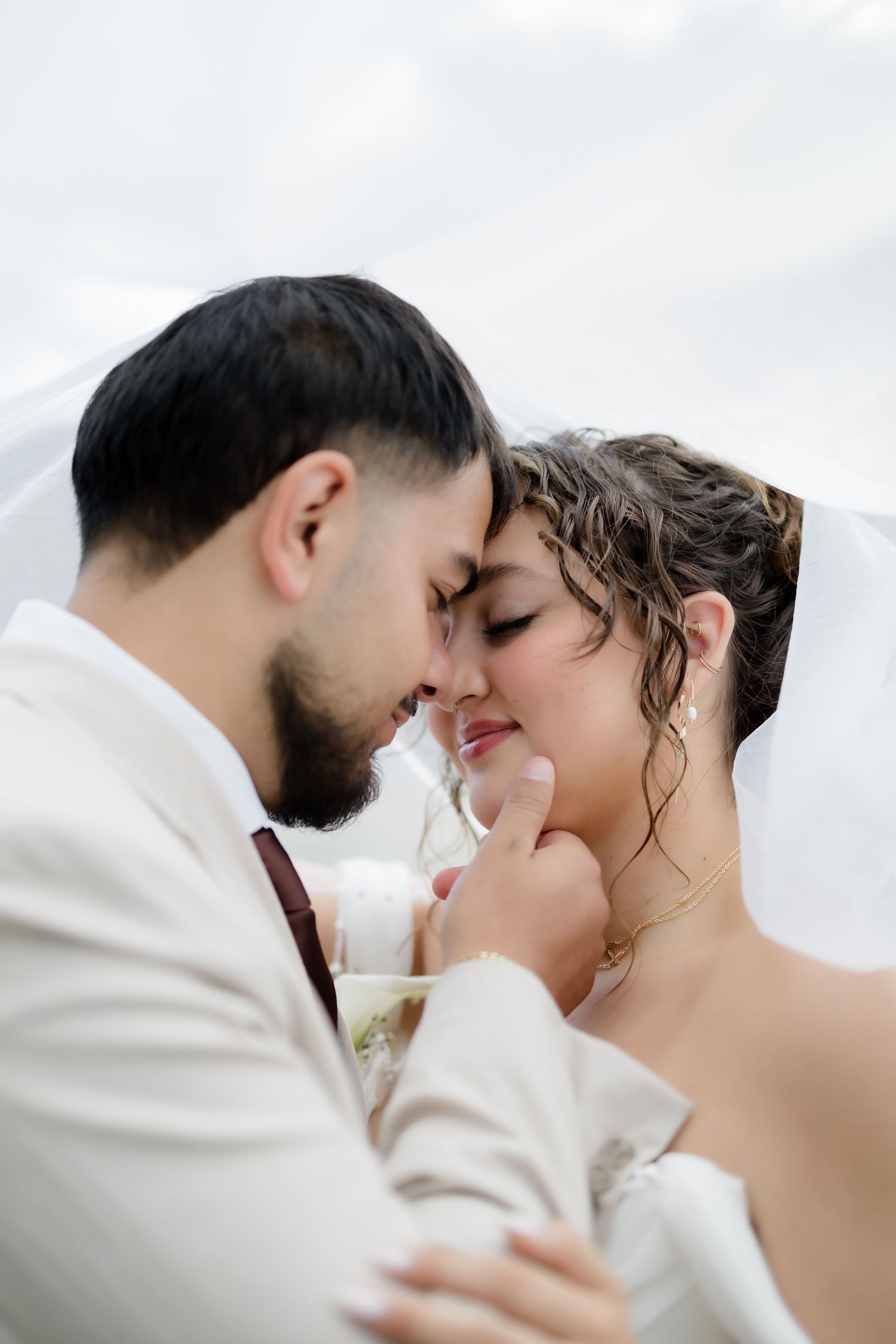 A bride and groom share an intimate forehead touch under a white veil on their wedding day.