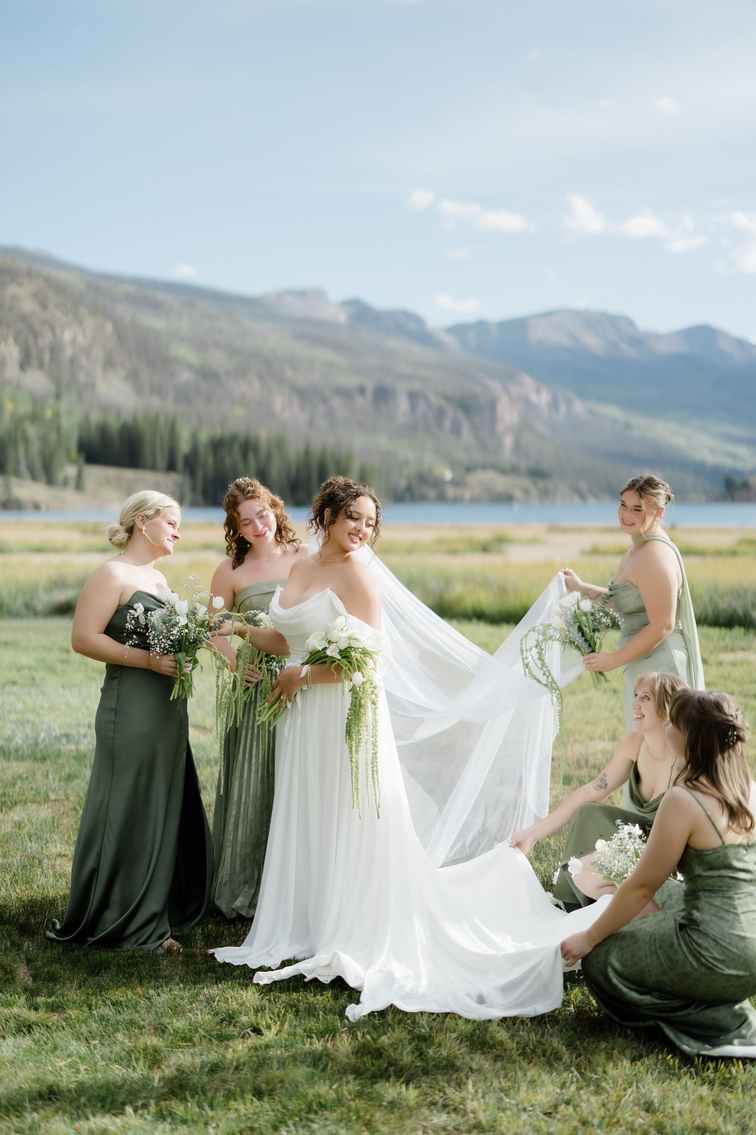 Bride in white gown with bridesmaids in green dresses adjusting veil, mountain lake backdrop.