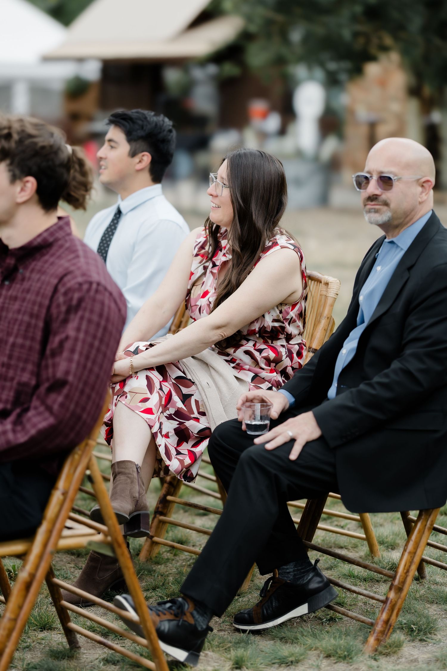 Guests seated in wooden chairs at an outdoor wedding ceremony, dressed in formal and semi-formal attire.