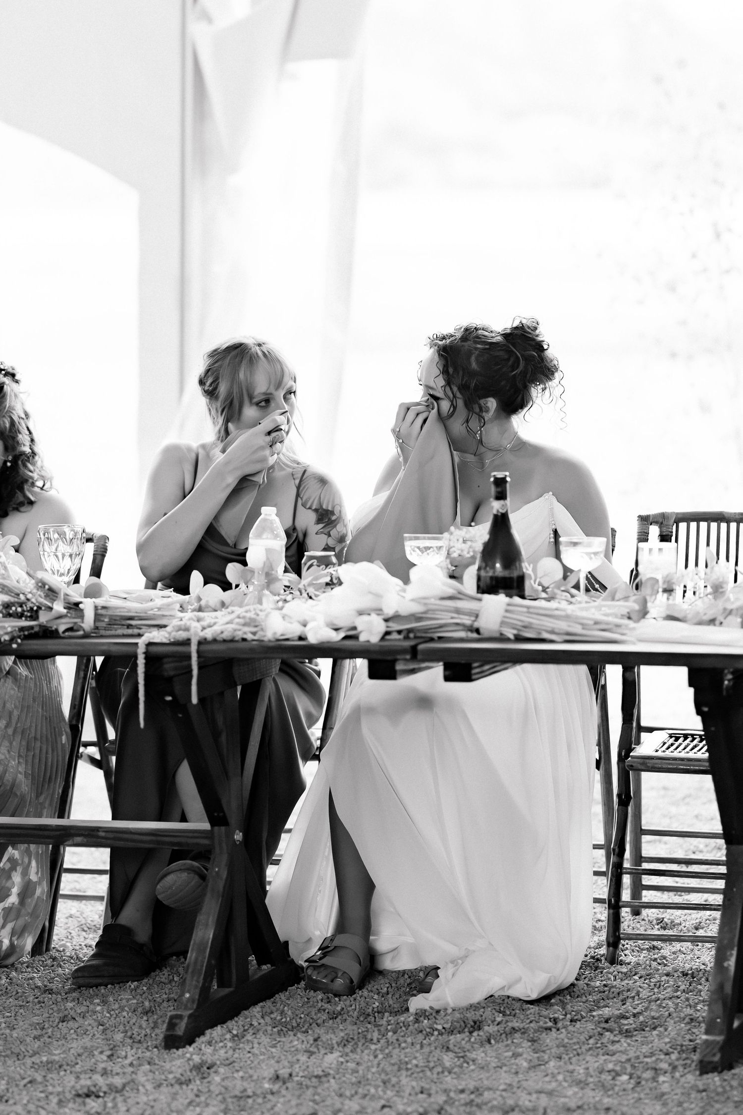 Black and white photo of bridesmaids crying at a wedding reception table with wine and floral decor.