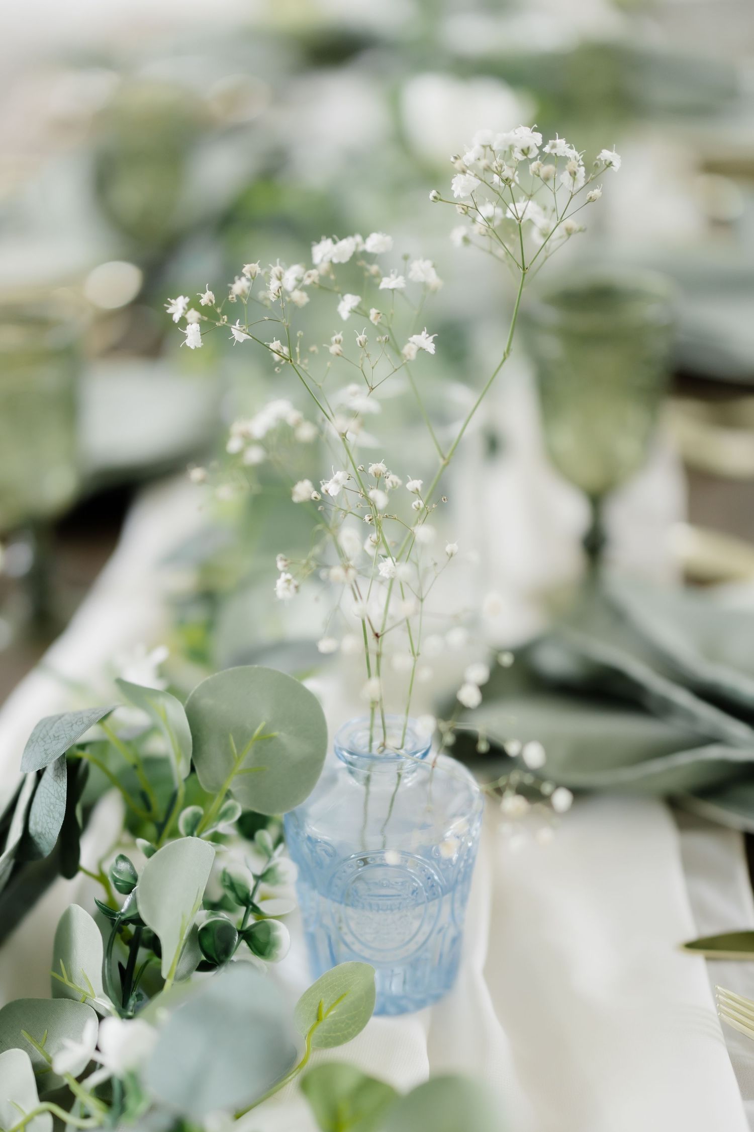 Elegant wedding table with baby's breath in blue vase, eucalyptus greenery, white linen, and sage green glassware.