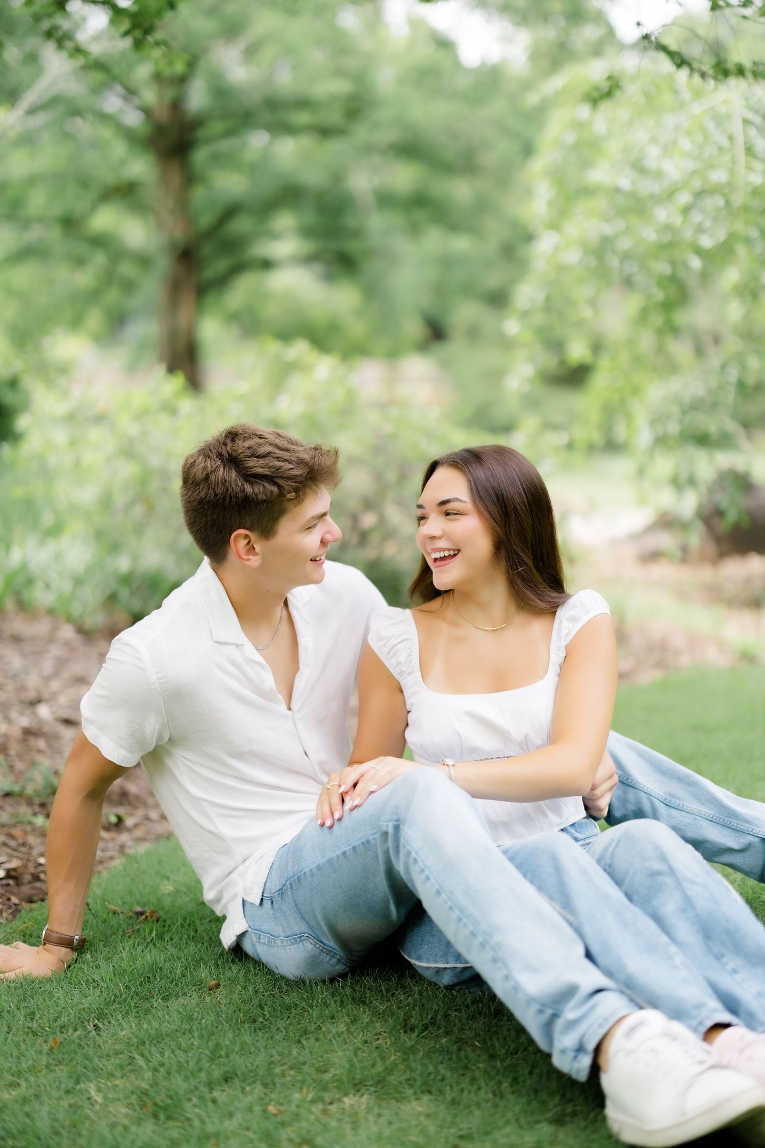 A young couple sitting on green grass in a park, laughing and enjoying each other's company on a sunny day.