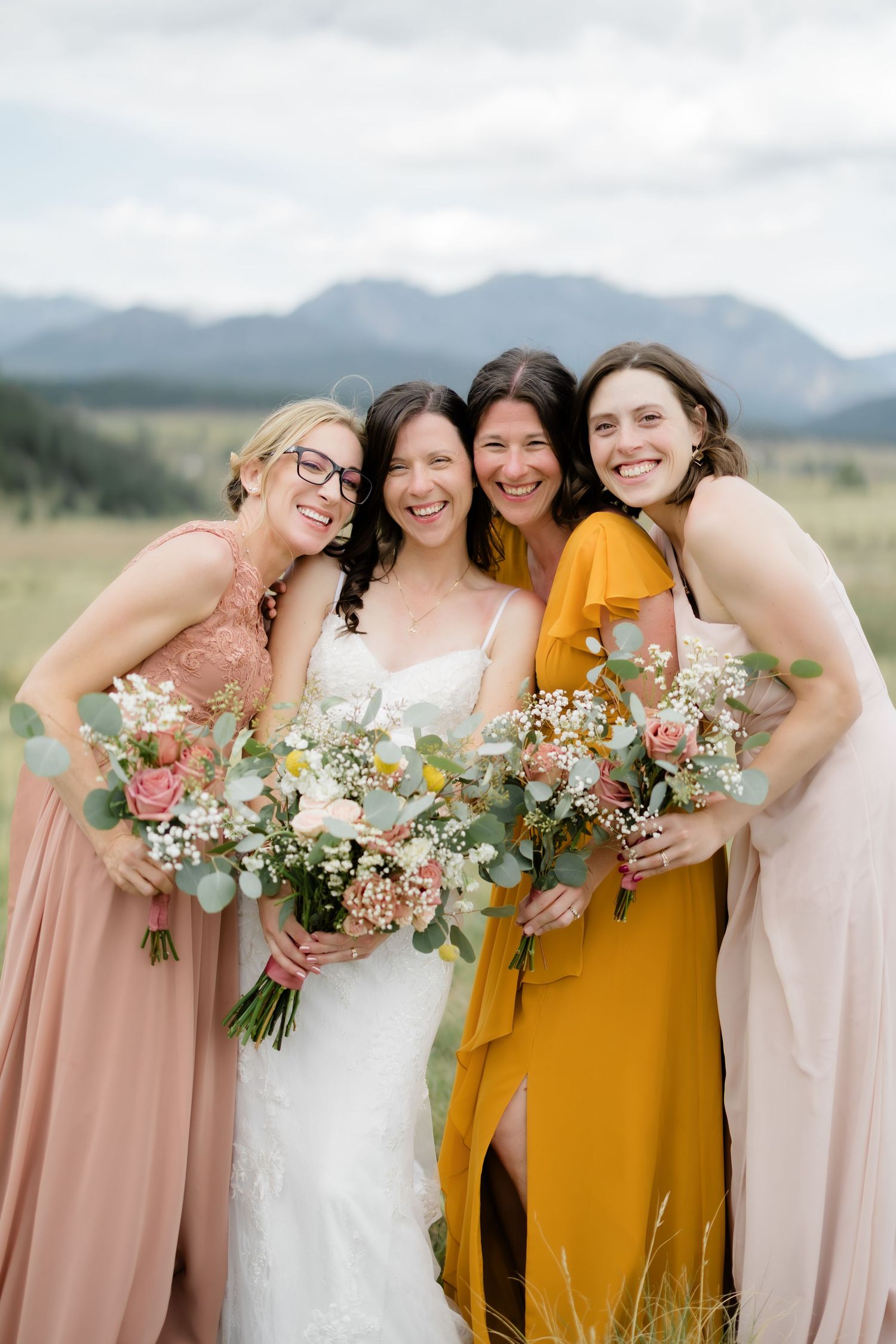 Bride in white gown with bridesmaids in pink and mustard dresses holding floral bouquets outdoors with mountain backdrop.