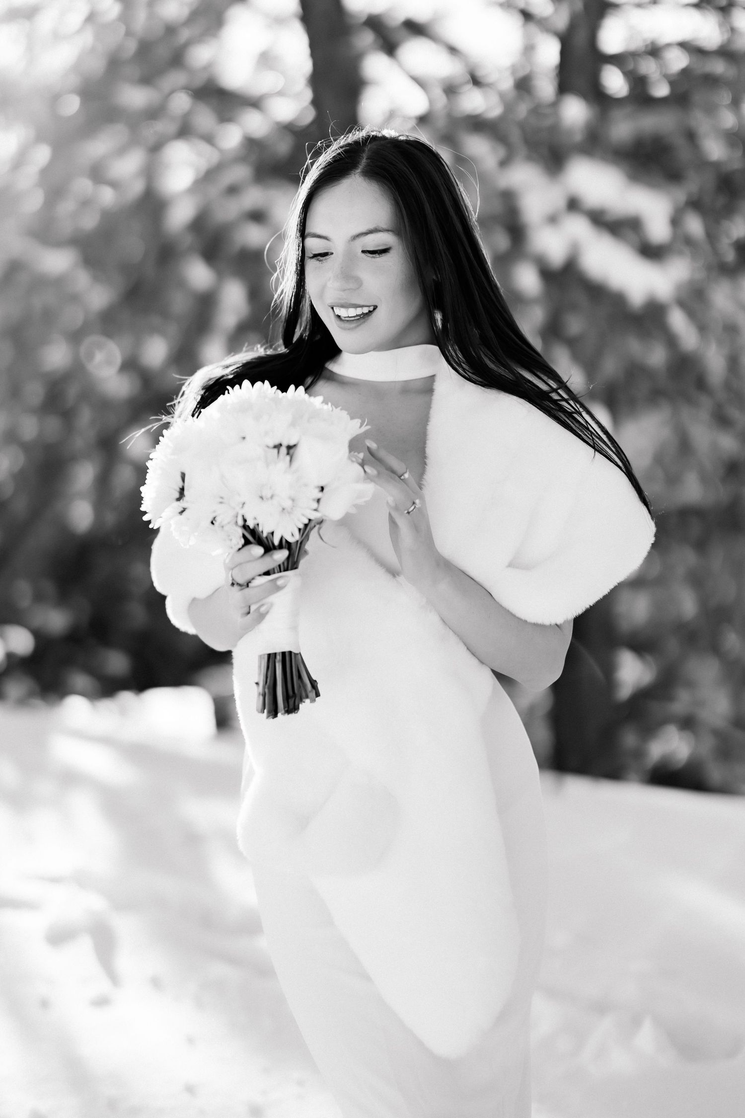 Black and white photo of smiling bride in fur wrap holding white bouquet in a snowy winter setting.