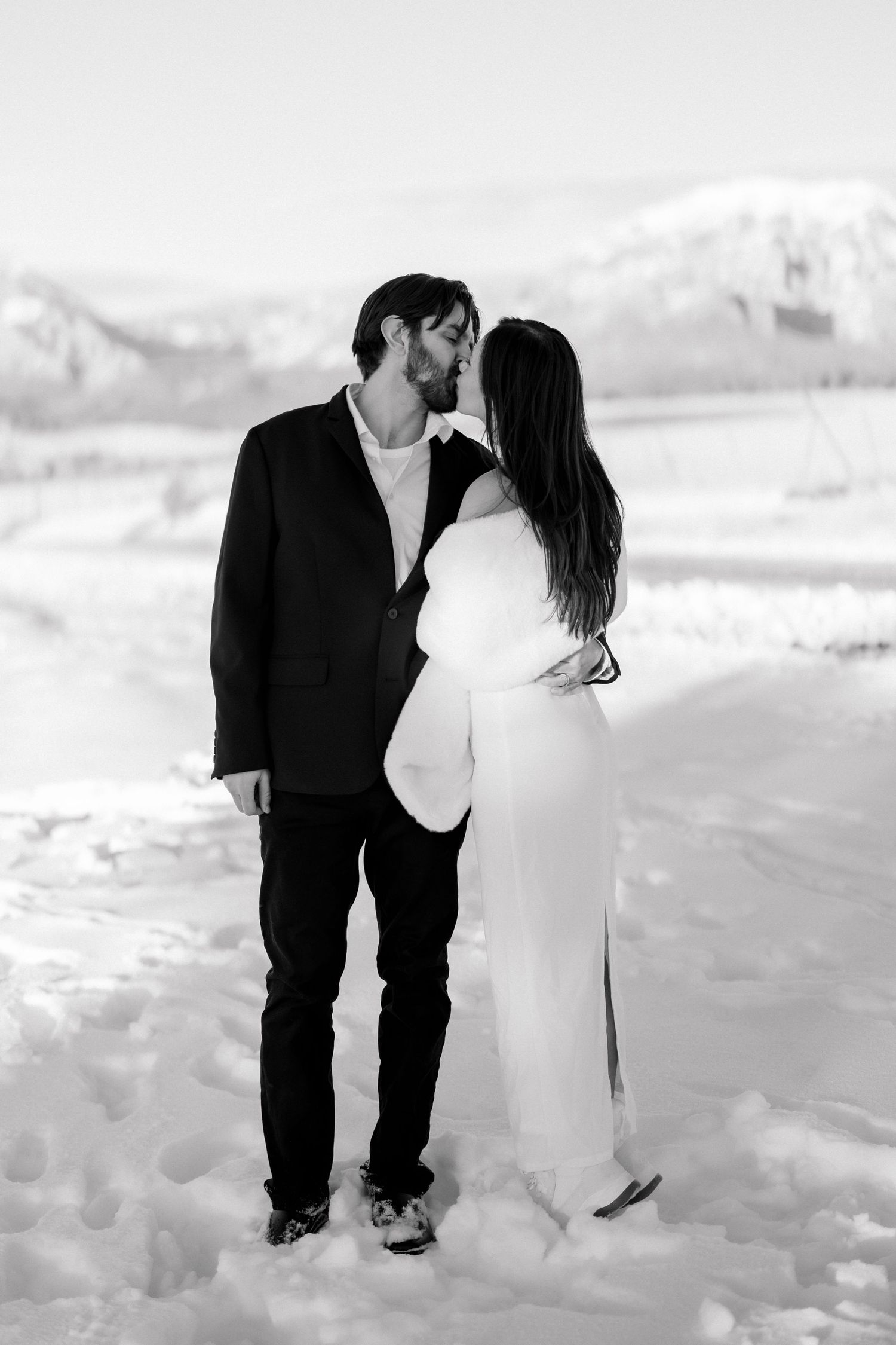 Bride and groom share a tender kiss standing in deep snow with a vast snowy mountain landscape behind them.
