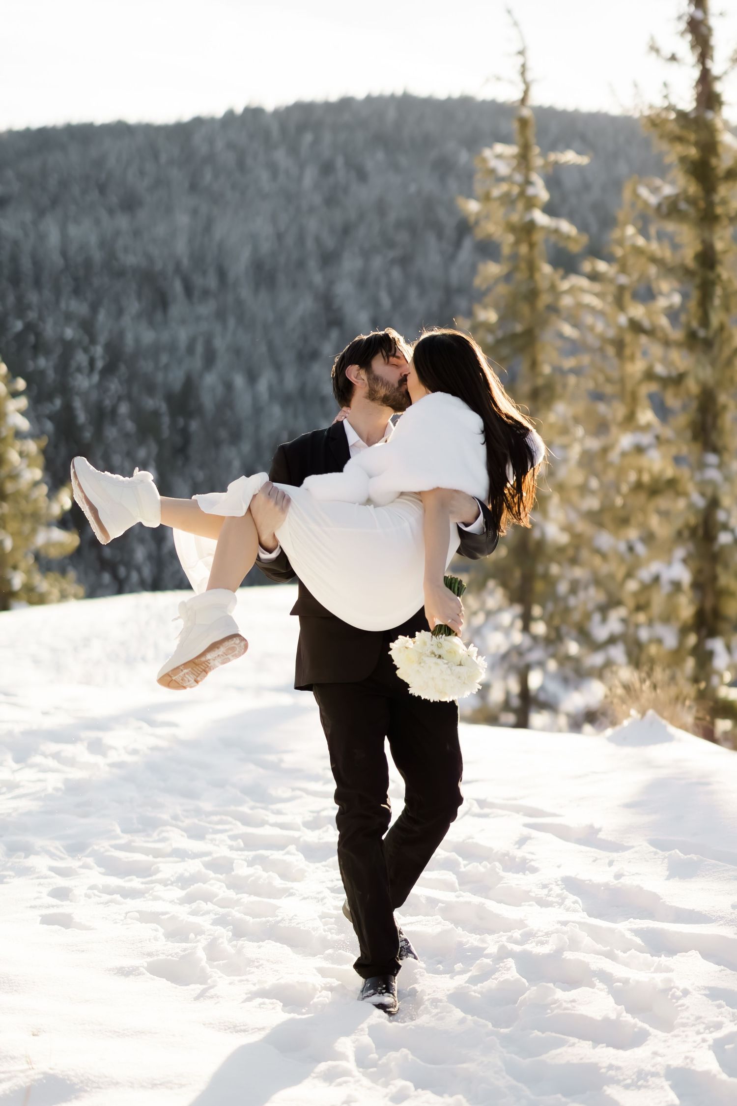 Groom carrying his bride in a white dress and booties through a bright snowy winter landscape.