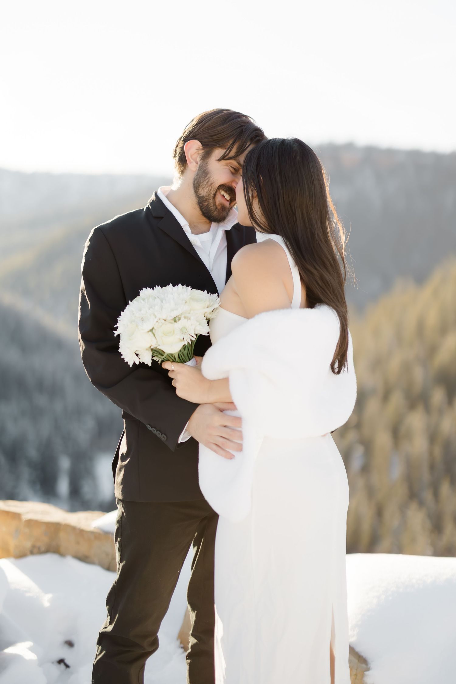 A couple shares a romantic kiss in winter wedding attire, with snowy mountain scenery in the background.