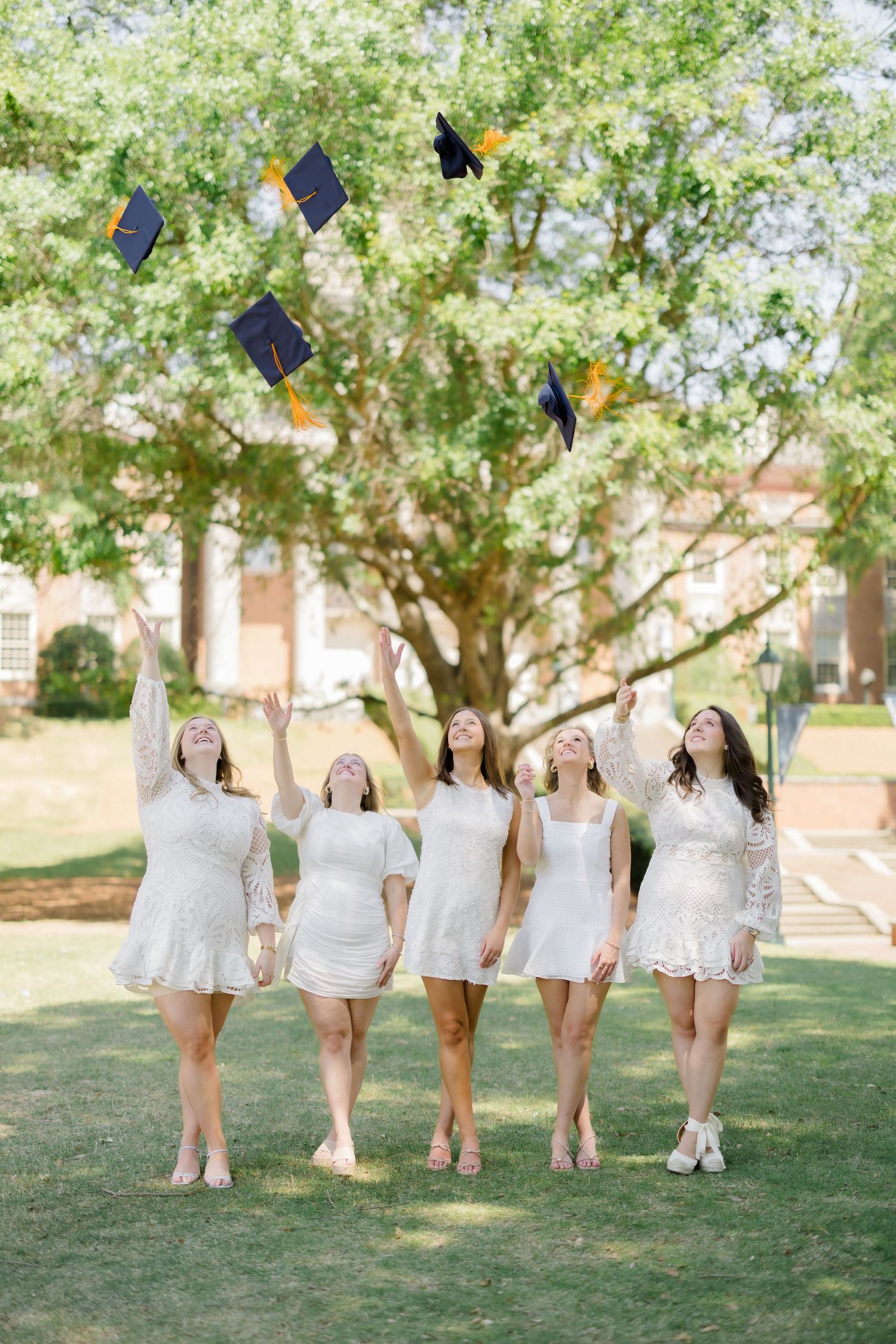 Five women in white dresses toss graduation caps in the air outdoors, celebrating their academic achievement.