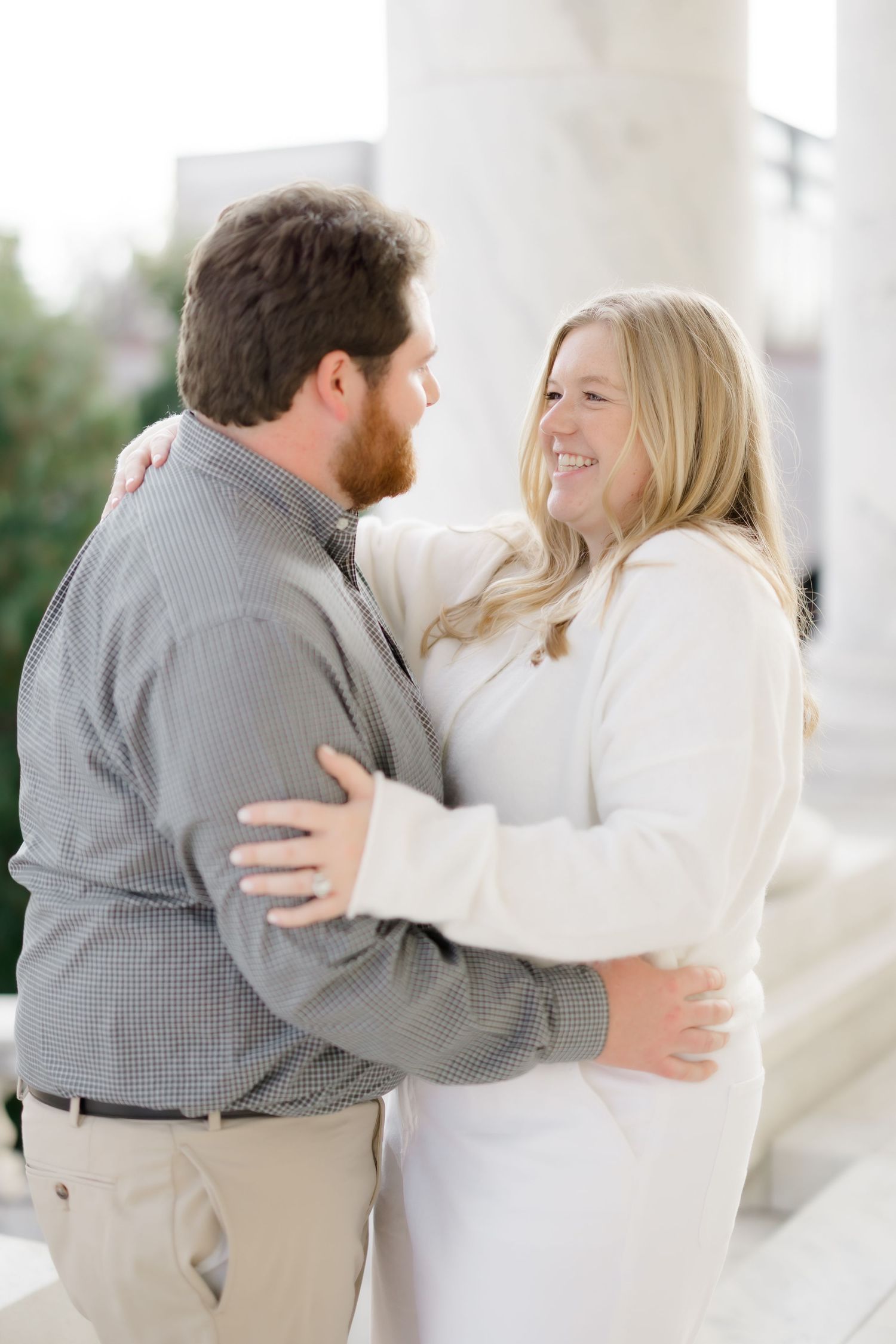 Couple embraces and smiles at each other near marble columns, woman in white outfit, man in gray shirt.