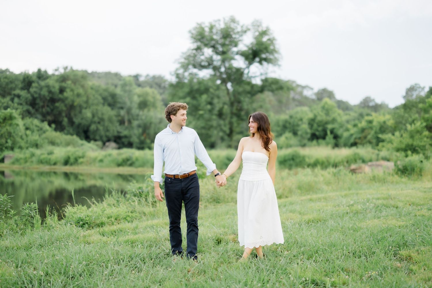 A couple holds hands walking through a lush green field near a pond, woman in white dress, man in blue shirt.