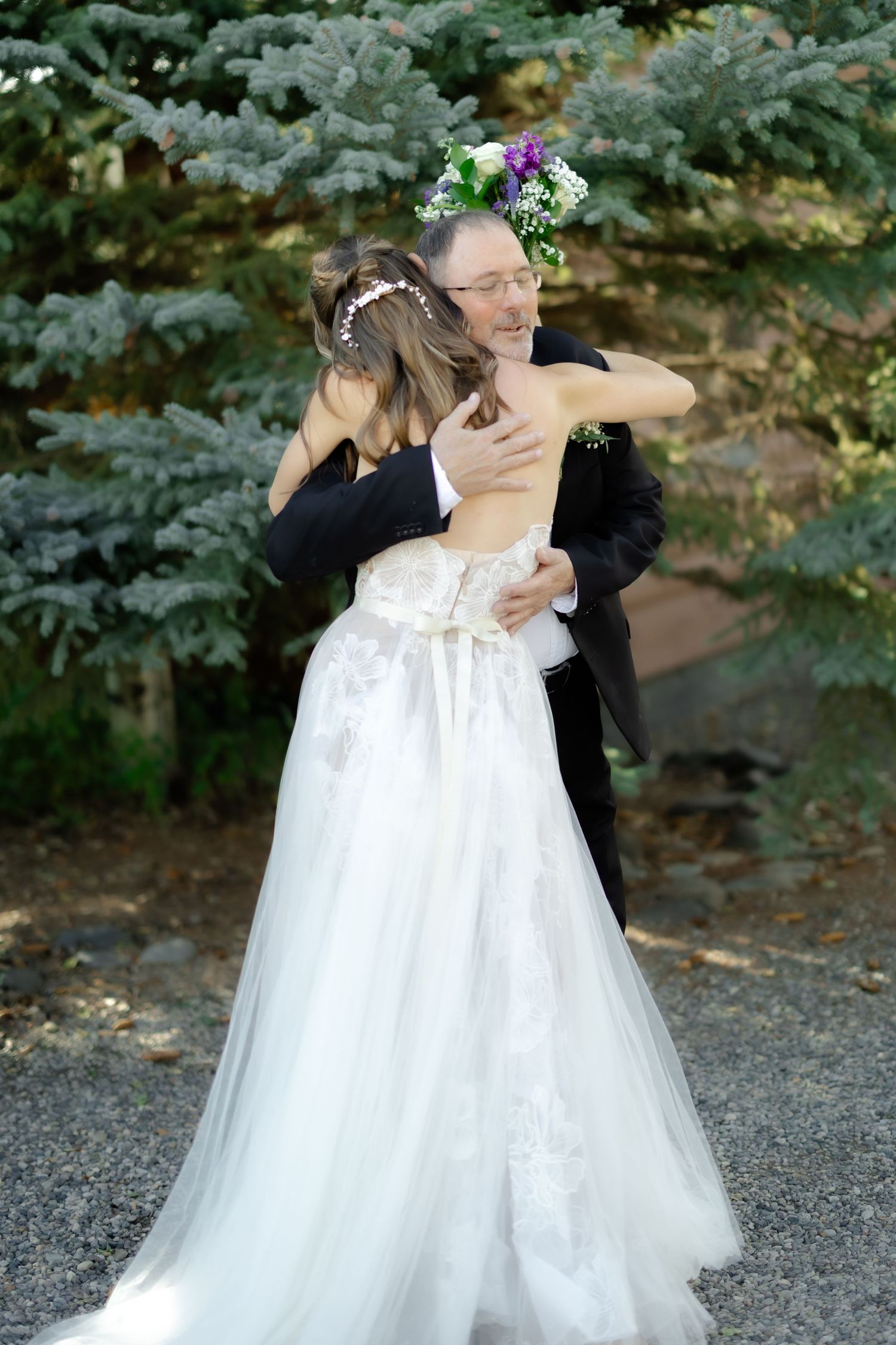 Bride in white lace gown hugging man in black suit outdoors near evergreen trees at wedding celebration.