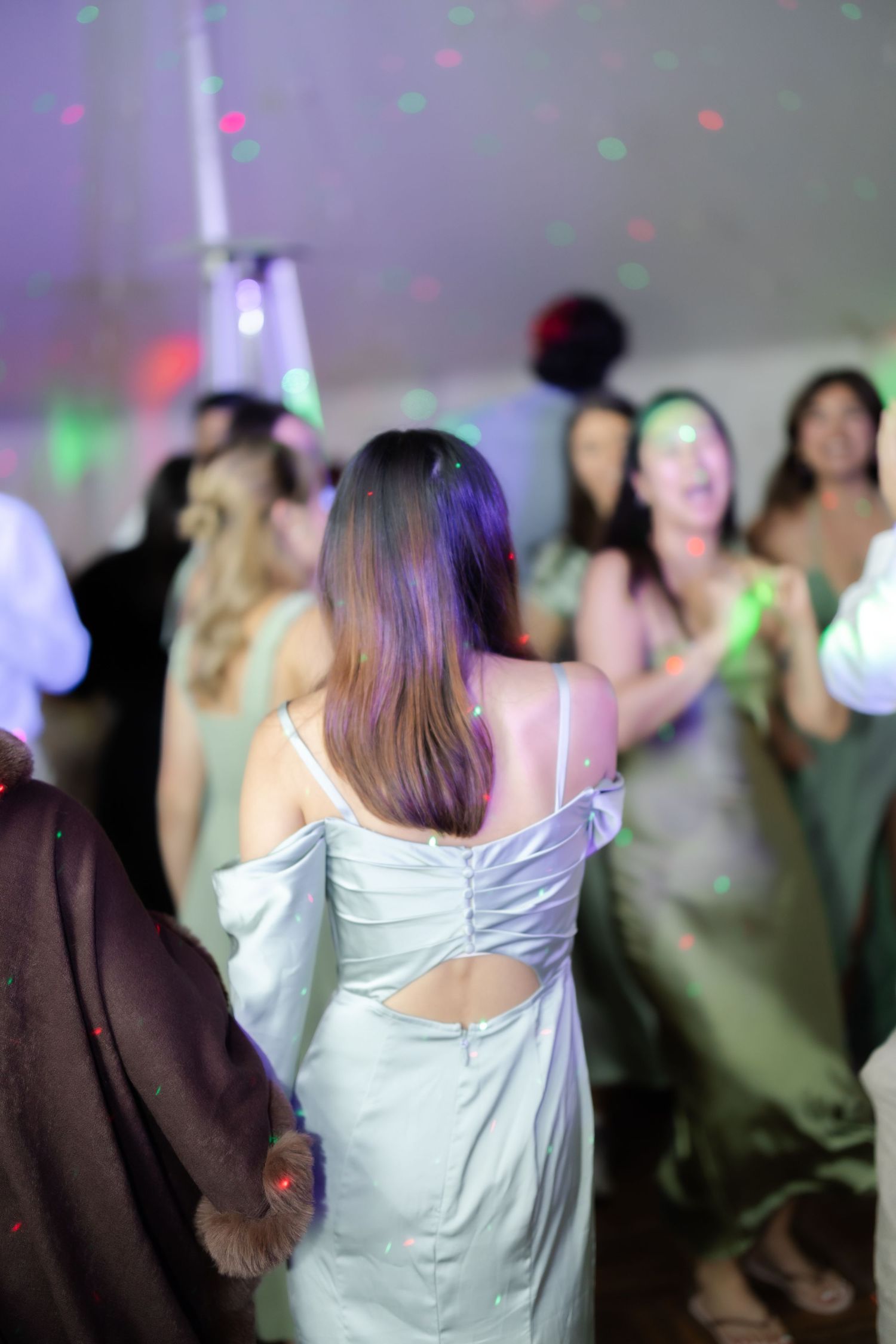 Women in elegant sage green and white dresses dancing at a formal event with colorful party lights.