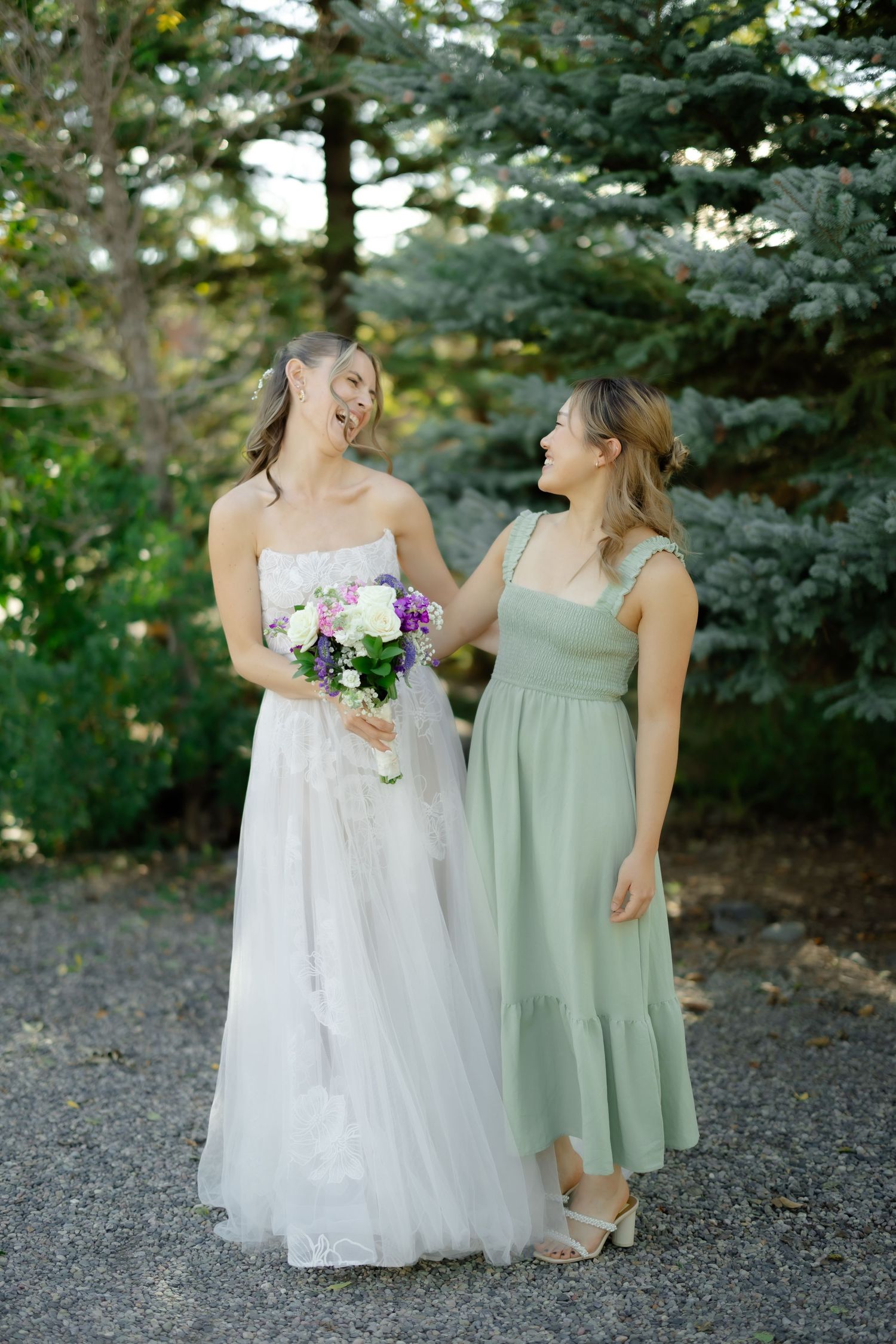 Bride in white gown holding bouquet laughs with bridesmaid in sage green dress outdoors near evergreen trees.