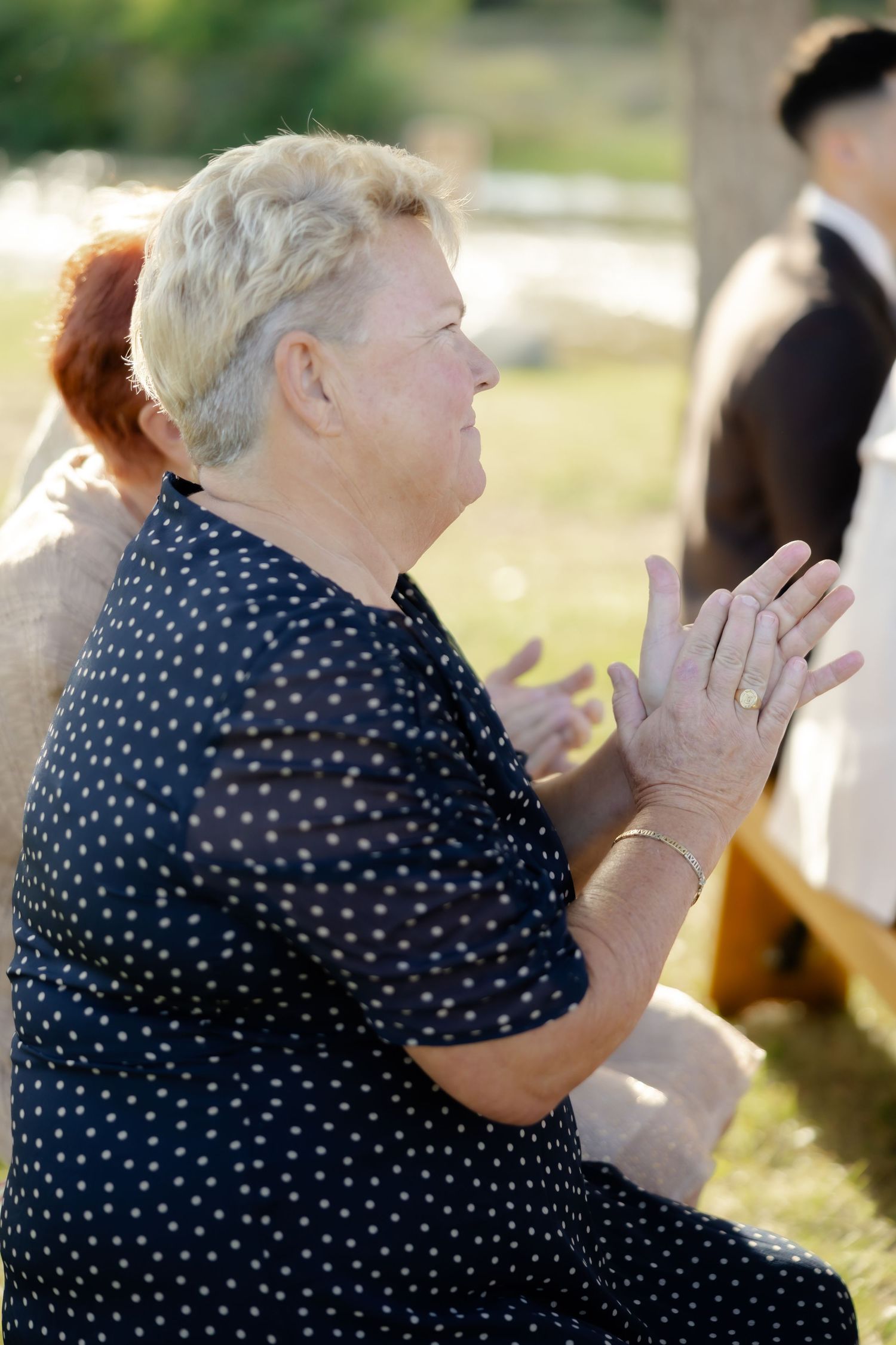 Elderly woman in polka dot dress clapping hands outdoors at a sunny gathering or wedding ceremony.