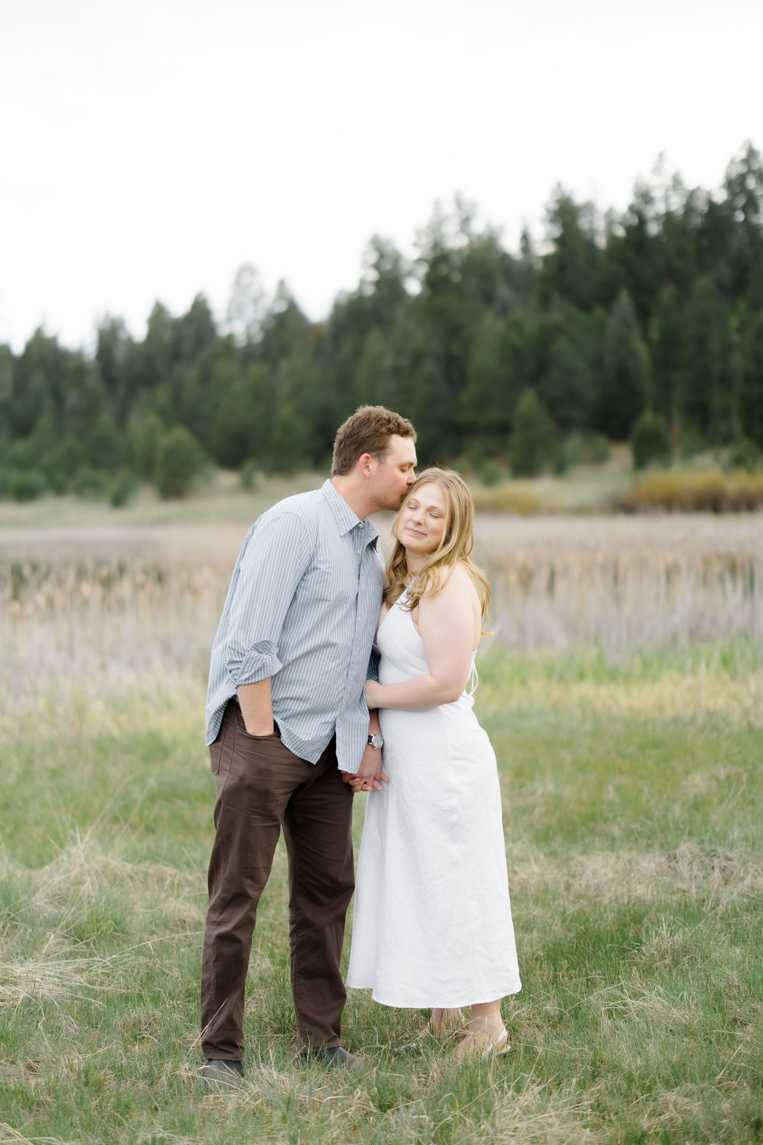 Man kissing woman on cheek while embracing in scenic meadow with pine forest backdrop.