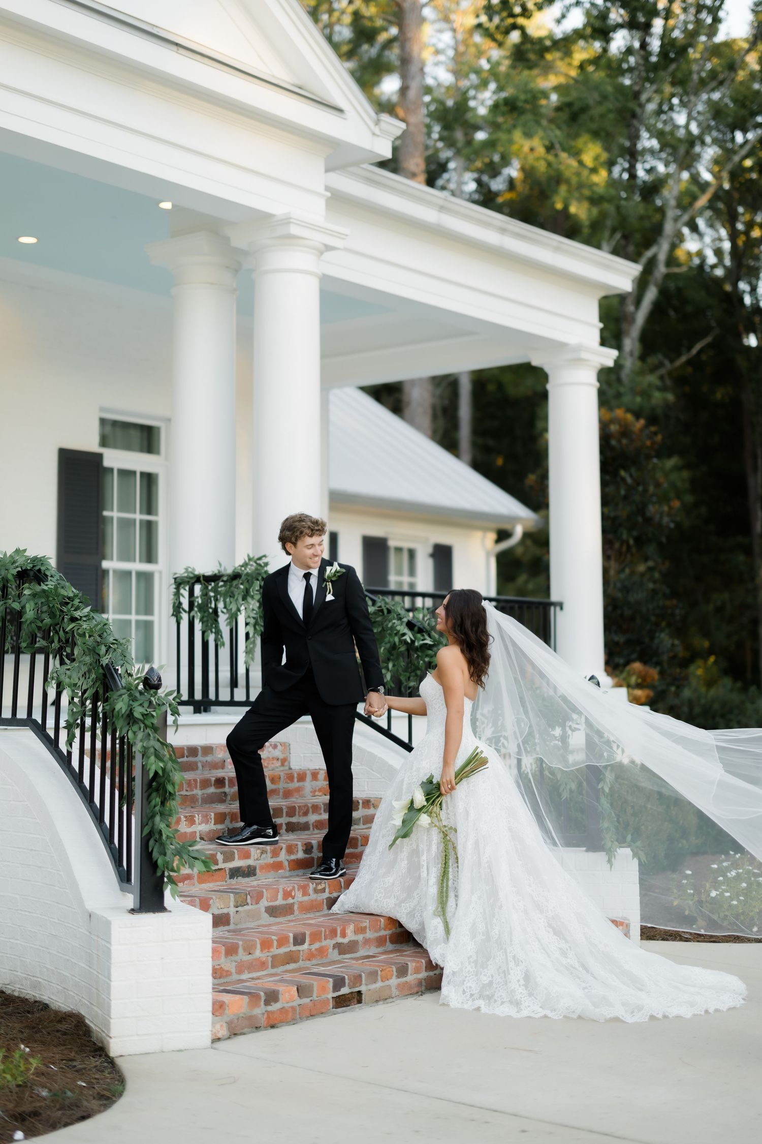 Bride in a lace wedding gown with a flowing veil holds hands with a groom in a black suit on brick steps by a white columned porch.