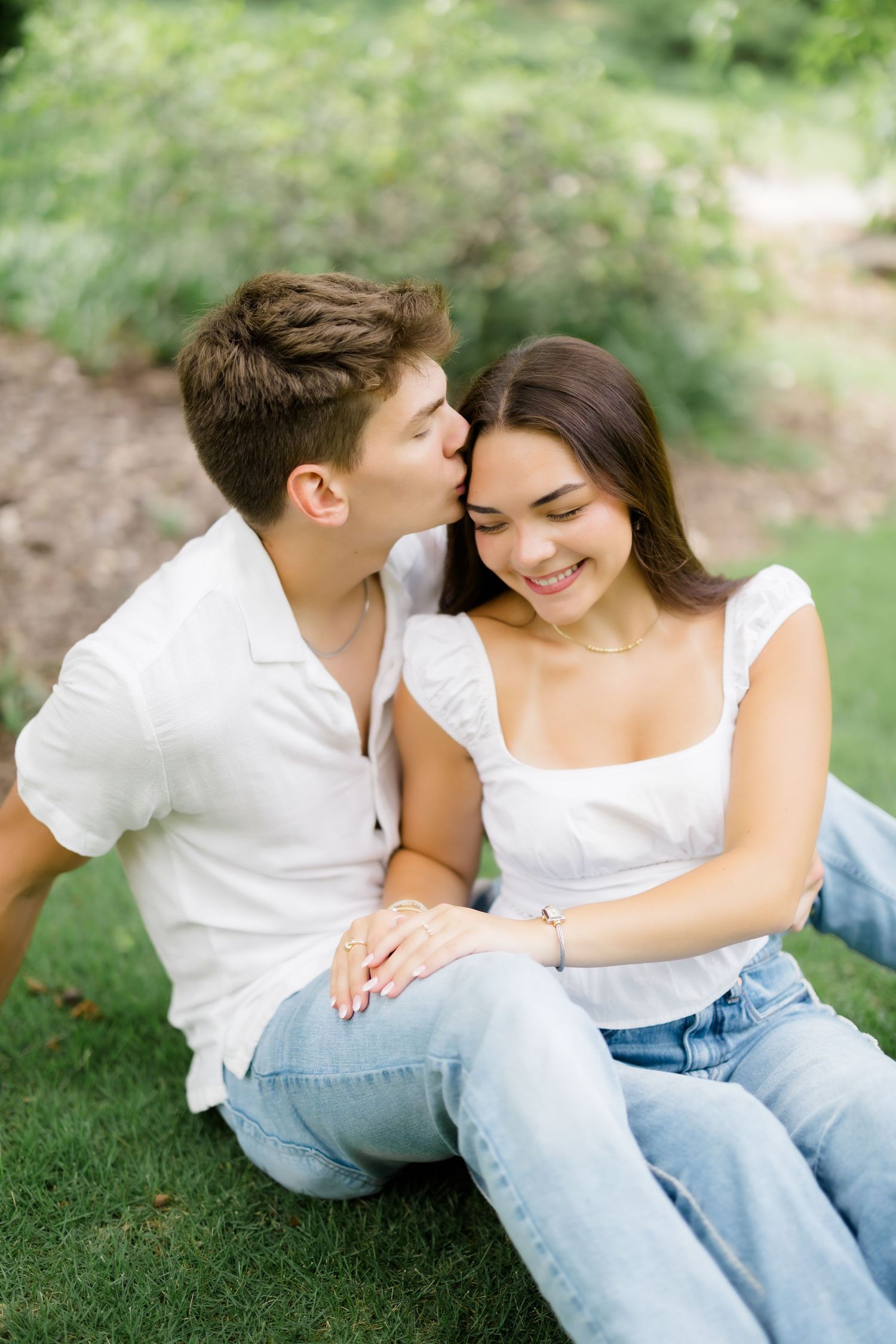 Young couple sitting on grass, man kissing woman on temple as she smiles, both in white and jeans.