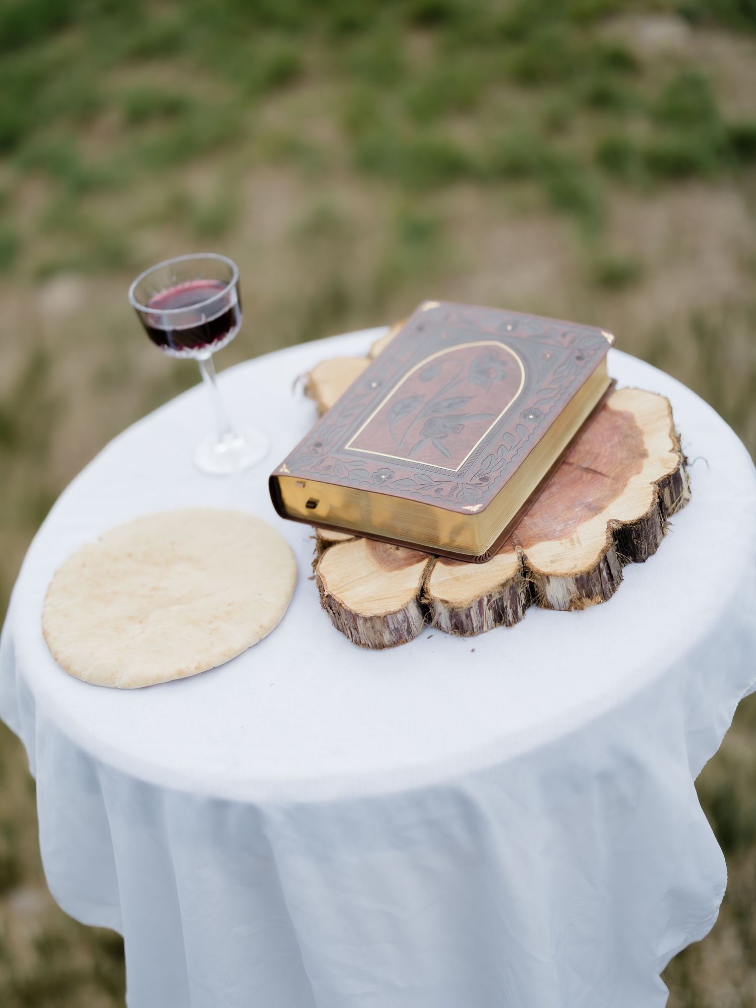 Rustic wedding communion table with Bible on wood slice, red wine glass, and bread on white linen.