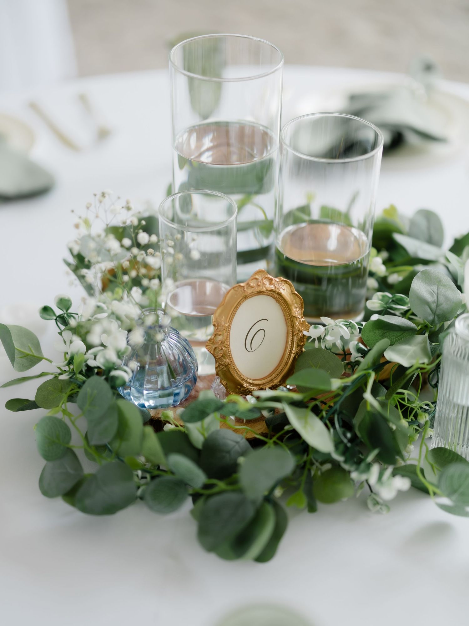Elegant wedding centerpiece with eucalyptus greenery wreath, glass cylinders, and a gold oval table number frame.