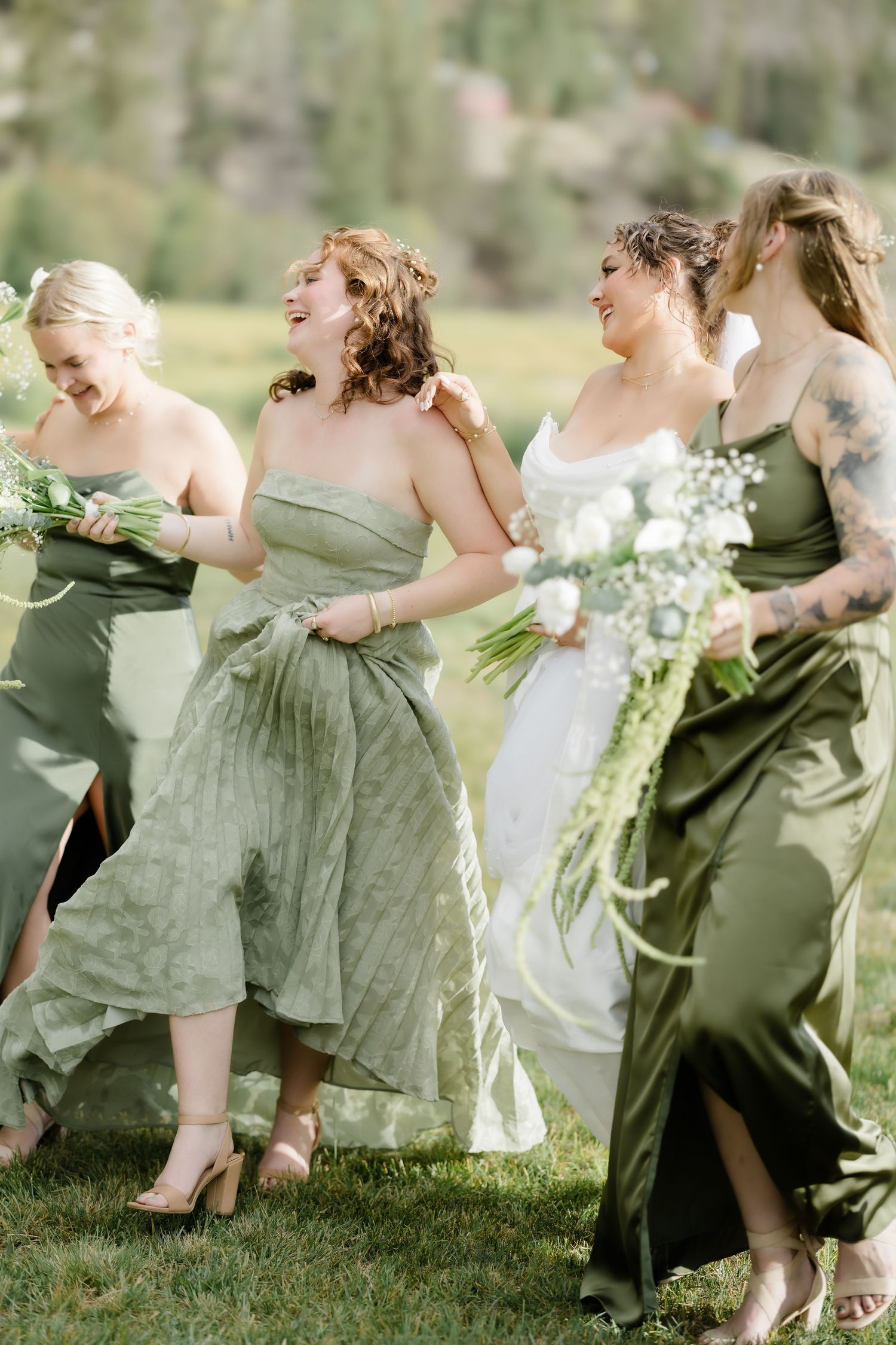 Bridesmaids in sage green dresses walk laughing with bride in white gown holding bouquets outdoors.