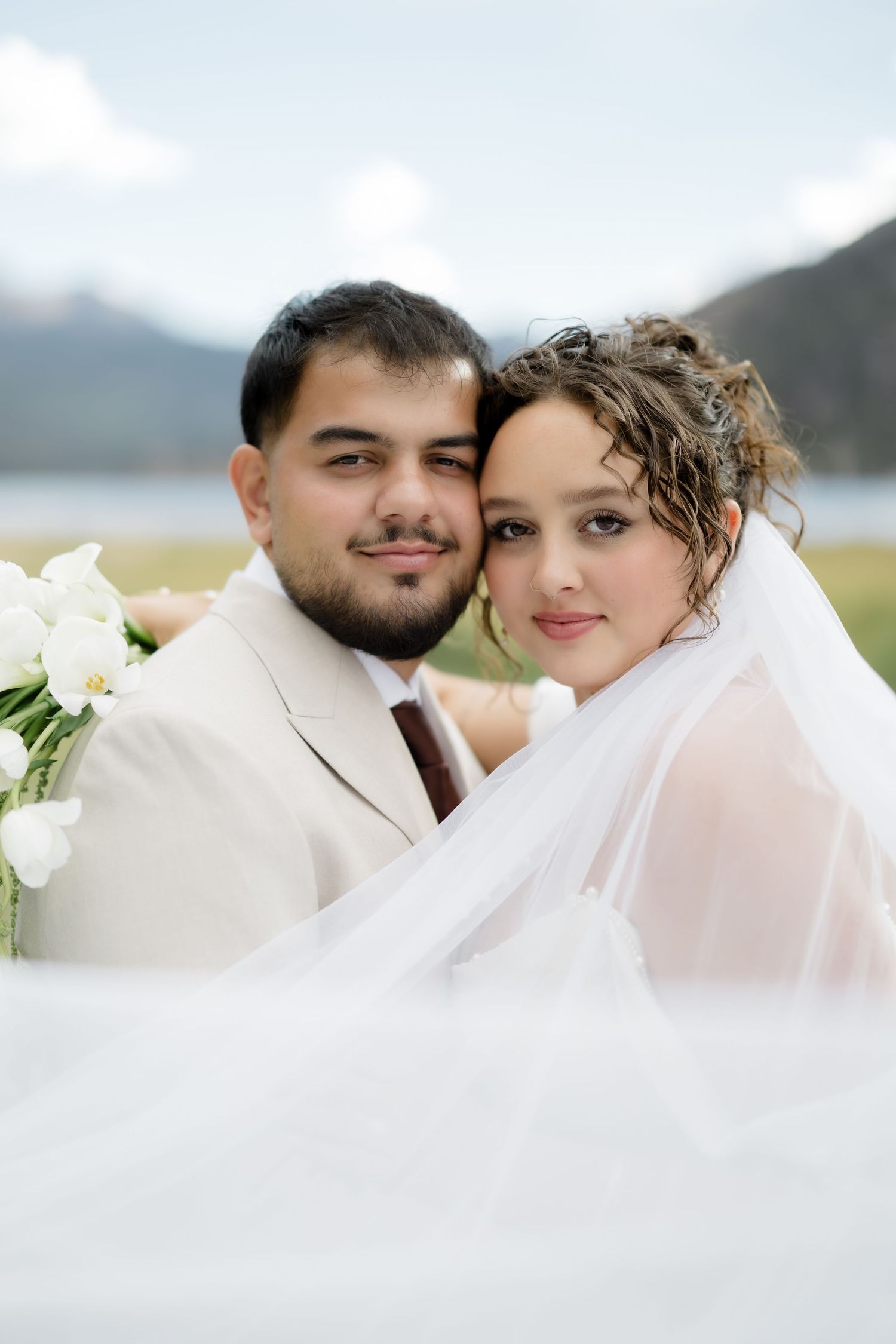 A bride and groom pose together outdoors near a lake and mountains, the bride wearing a veil and holding white calla lilies.