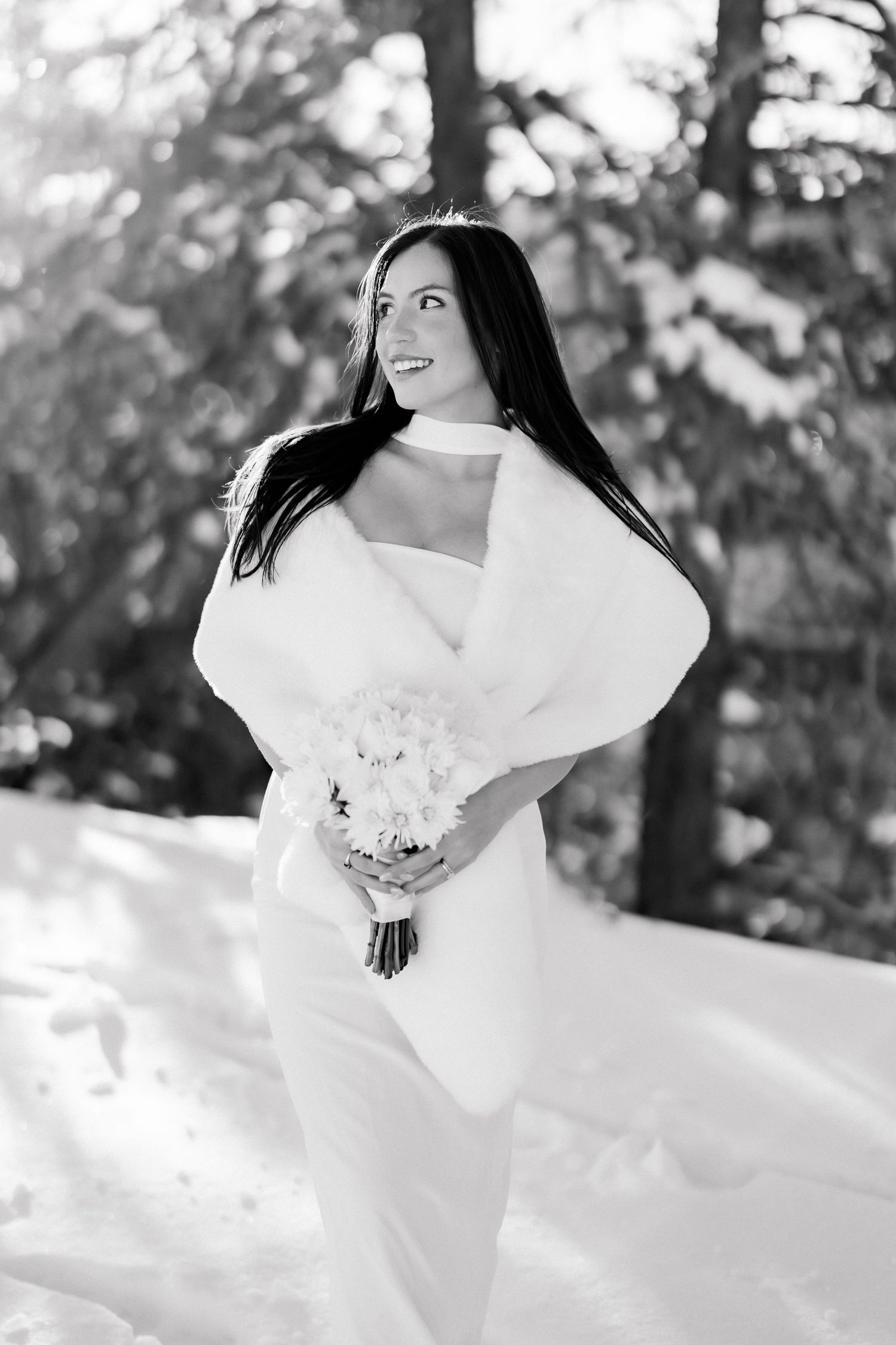 Smiling bride in white dress and fur wrap holds bouquet in snowy winter forest, black and white photo.