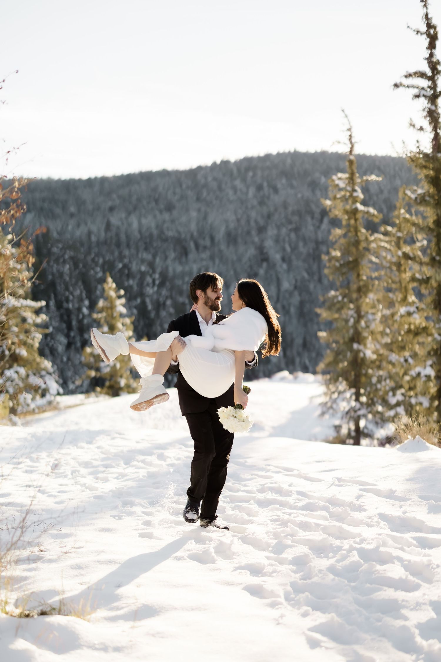 Groom lifting bride in white wedding dress in his arms on a snowy mountain trail surrounded by pines.