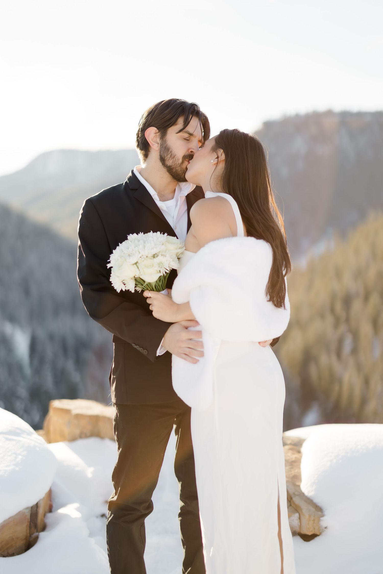 Bride and groom kissing in snowy mountain setting, bride in white dress holding bouquet, groom in black suit.