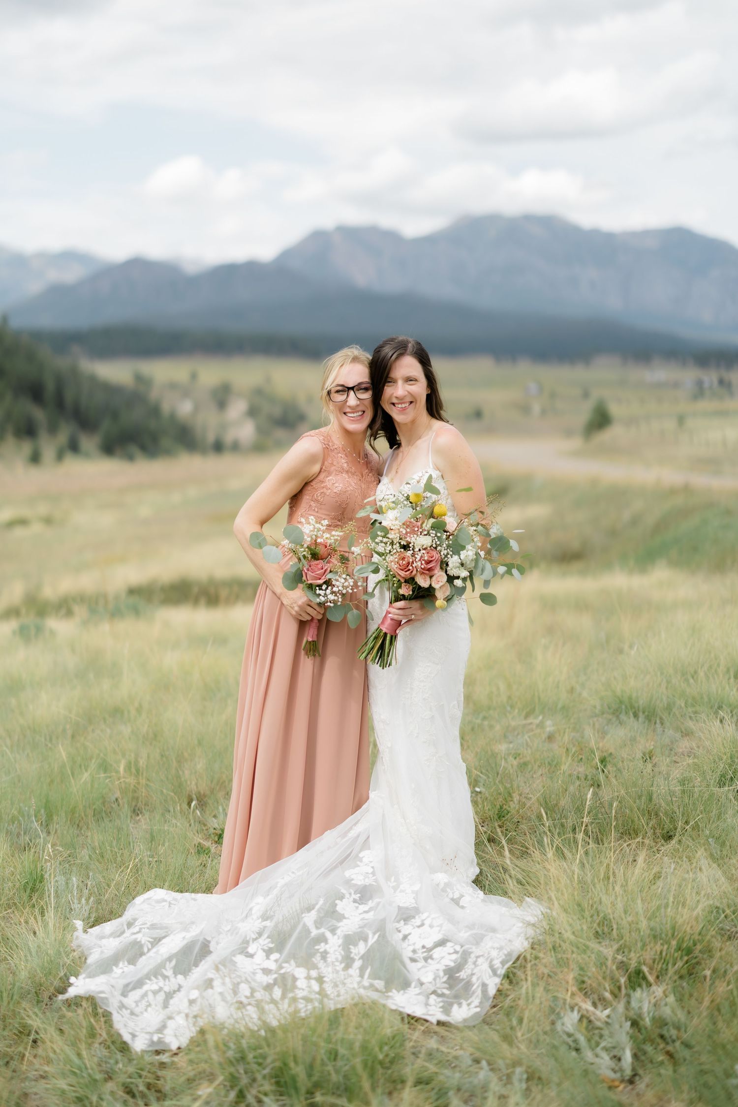 Bride in white lace gown and bridesmaid in dusty rose dress holding bouquets with mountain meadow backdrop.