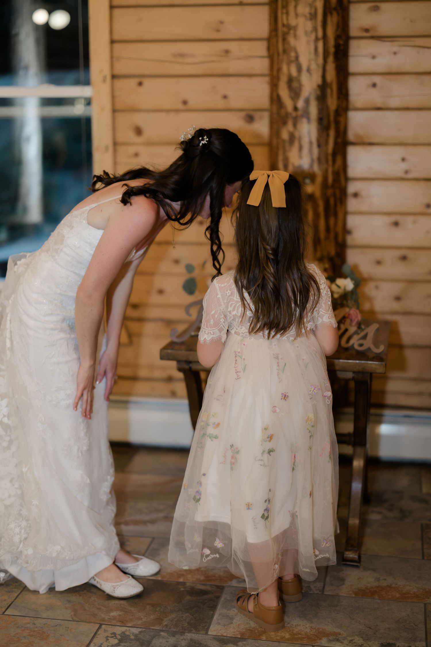 Bride in white lace gown bending down to speak with flower girl in floral dress with gold bow in rustic wood venue.