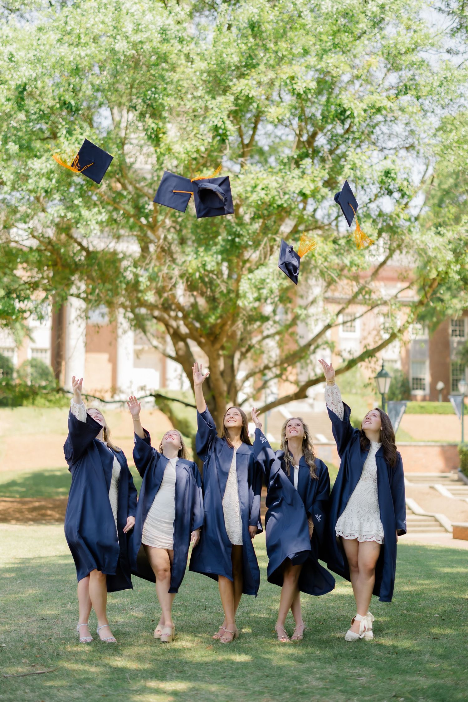 Five female graduates in navy gowns joyfully toss their mortarboard caps in the air outdoors.