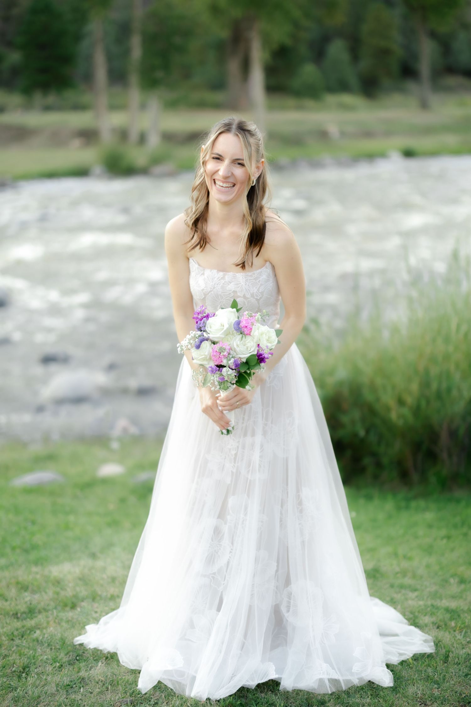 Smiling bride in a strapless lace tulle gown holding a colorful bouquet outdoors near a river.