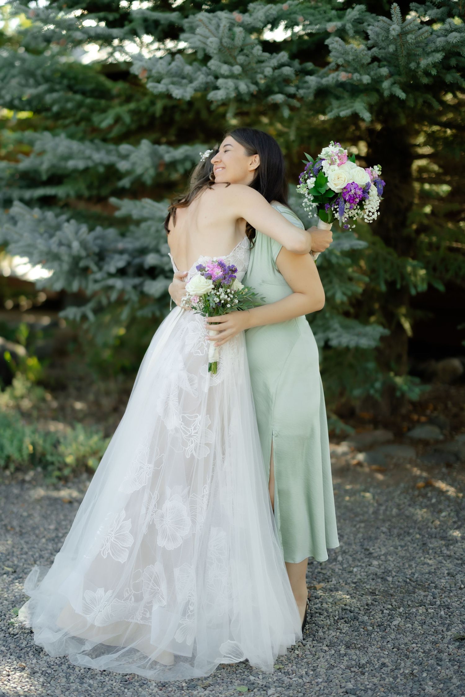 Bride in lace gown hugging bridesmaid in sage green dress, both holding bouquets near pine trees.